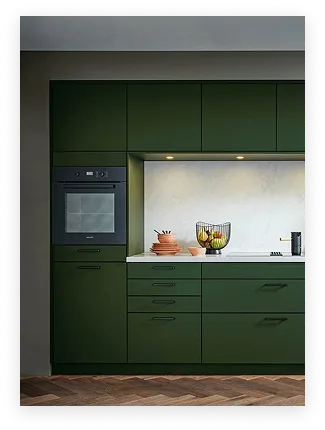 Cuisine moderne avec meubles verts foncés, four encastré, bol à fruits sur le plan de travail blanc et carrelage mural clair.