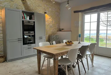 Cuisine avec table en bois rectangulaire, six chaises blanches, un panier de fruits au centre, et une baie vitrée donnant sur un paysage extérieur.