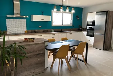 Cuisine moderne avec murs bleu canard, meubles blancs, table noire avec quatre chaises jaunes, et grand réfrigérateur en acier inoxydable.