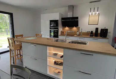 Cuisine moderne avec îlot central en bois clair, évier intégré, étagères éclairées, et grandes fenêtres donnant sur un jardin verdoyant.