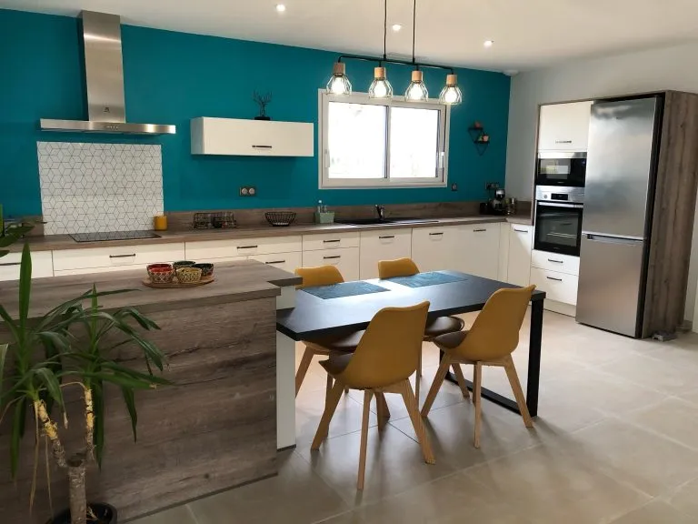 Cuisine moderne avec murs bleu turquoise, comptoirs en bois clair, table noire avec quatre chaises jaunes, et électroménagers en acier inoxydable.