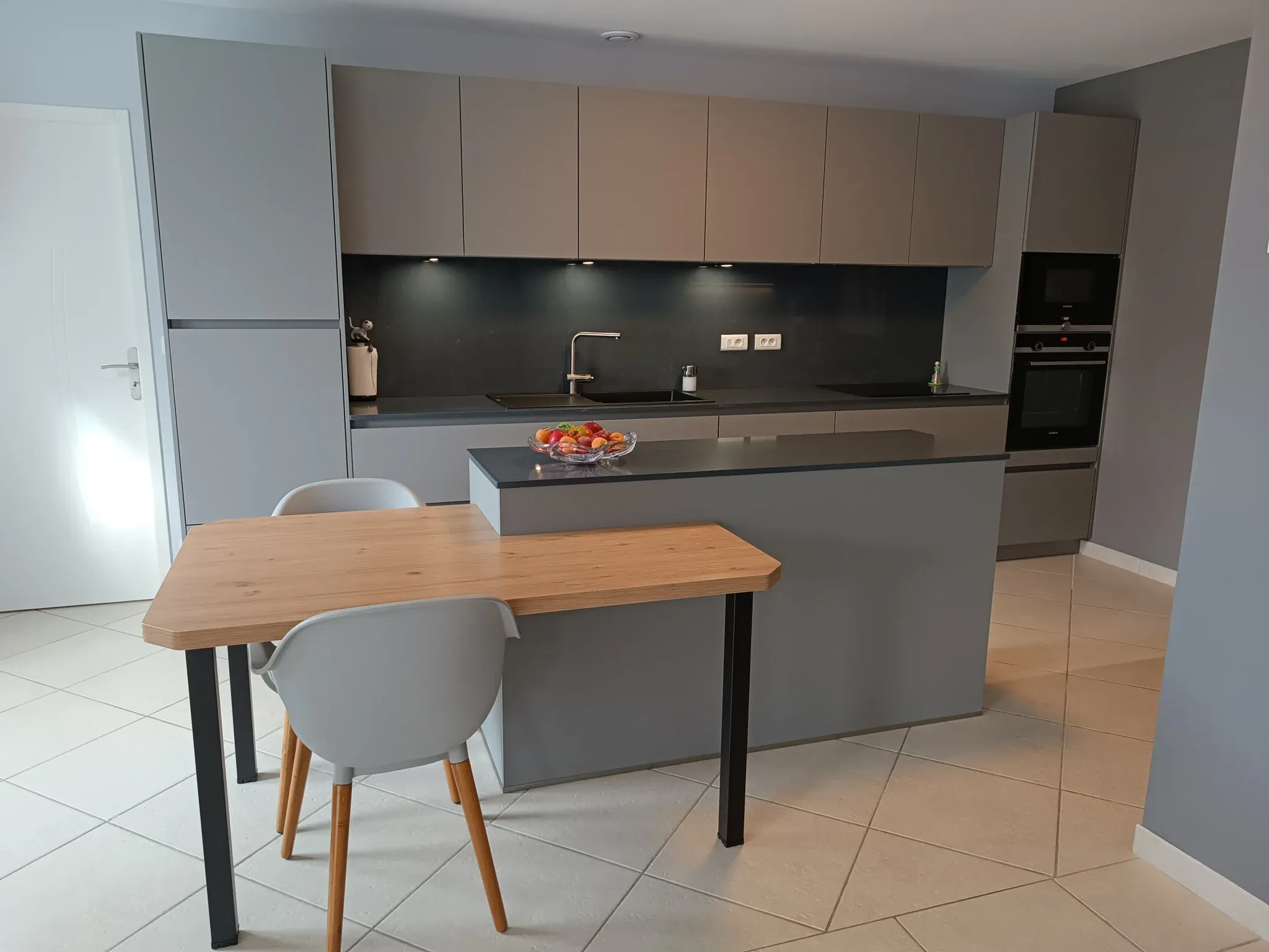 Cuisine moderne grise avec plan de travail noir, îlot central, table en bois clair avec deux chaises et bol de fruits.