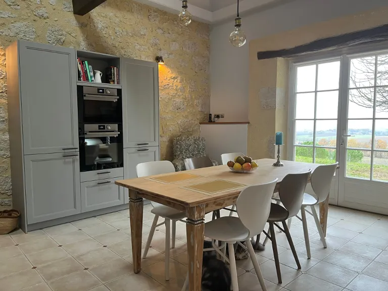 Cuisine avec une table en bois et six chaises, une grande baie vitrée donnant sur un paysage extérieur, et un mur en pierre avec un meuble de cuisine gris équipé d'appareils électroménagers.