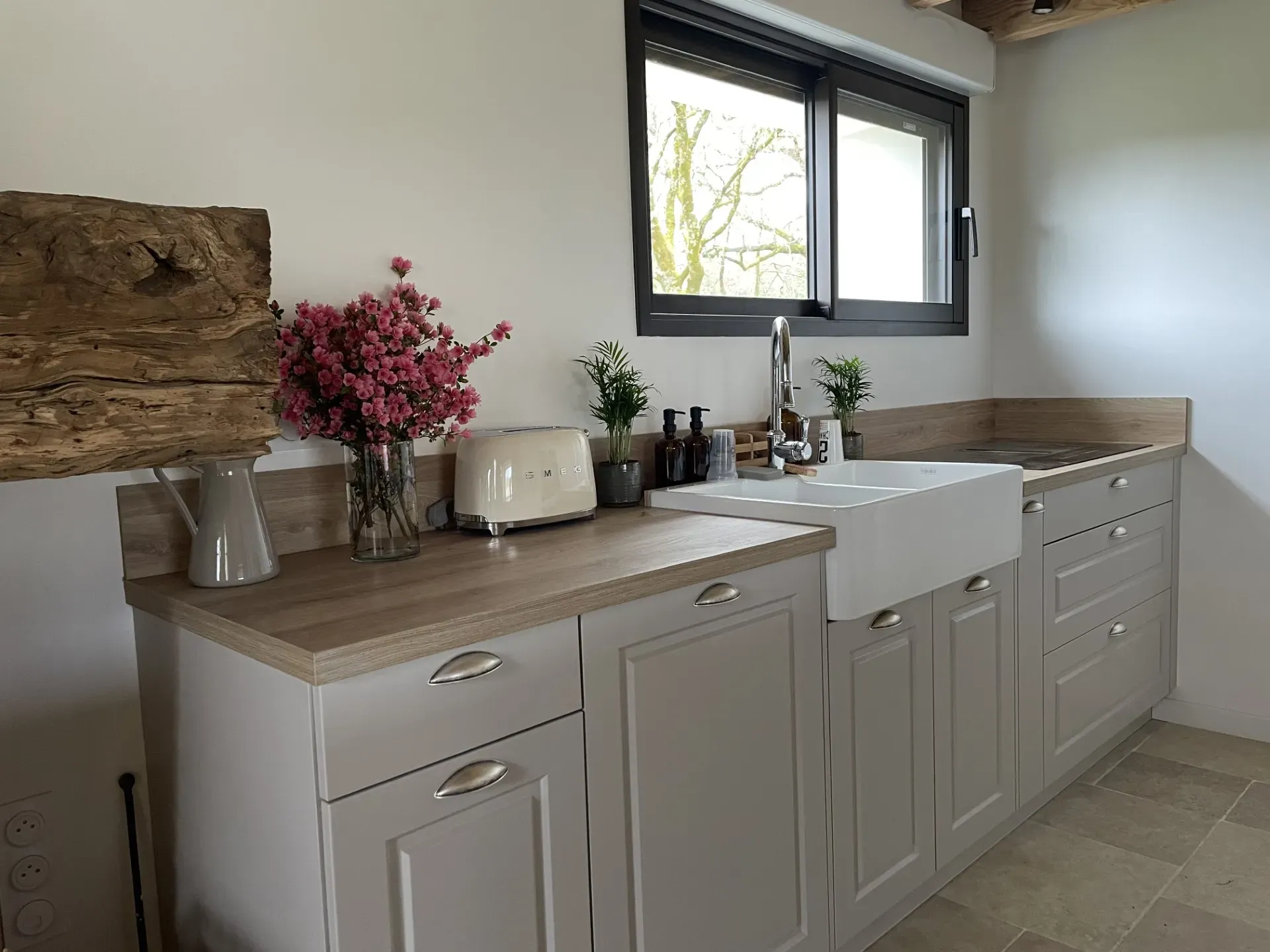 Cuisine moderne avec plan de travail en bois, évier blanc, grille-pain SMEG crème, plantes vertes et bouquet de fleurs roses sous une fenêtre.