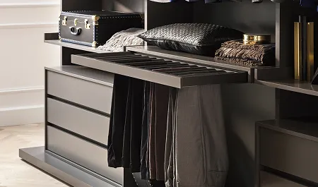 Dressing moderne avec des pantalons suspendus sous un tiroir coulissant, des coussins et une valise noire posés sur des étagères.