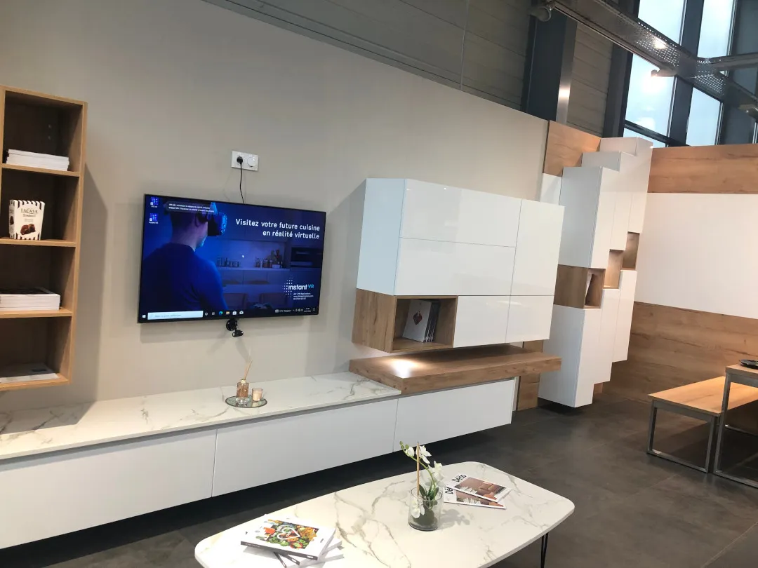 Salon moderne avec meubles blancs et en bois, une télévision murale affichant une publicité pour une visite de cuisine en réalité virtuelle, et une table basse en marbre avec un vase et des magazines.