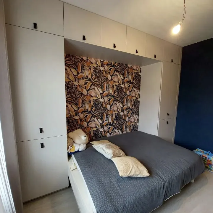 Chambre avec un grand lit couvert d'une couette grise et de deux coussins beiges, avec un mur décoré de papier peint à motifs feuilles, entouré par des placards blancs encadrant le lit.