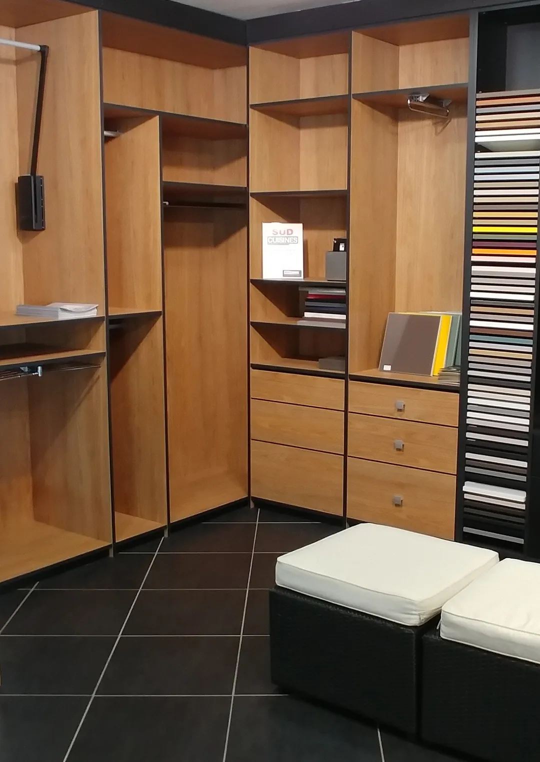 Coin d'un placard en bois avec étagères, tiroirs et échantillons de couleurs, au-dessus de deux poufs noirs avec coussins blancs.