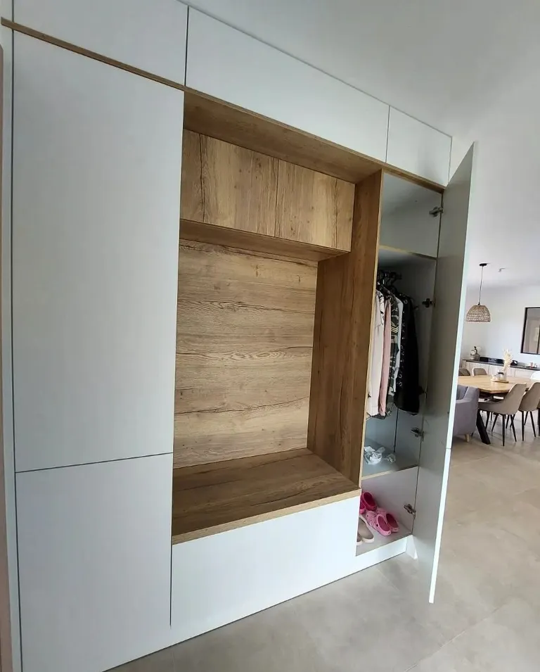 Placard encastré avec finition bois clair et portes blanches, avec un compartiment ouvert comportant un banc en bois, une porte ouverte montrant des vêtements suspendus et des chaussures roses sur le sol.