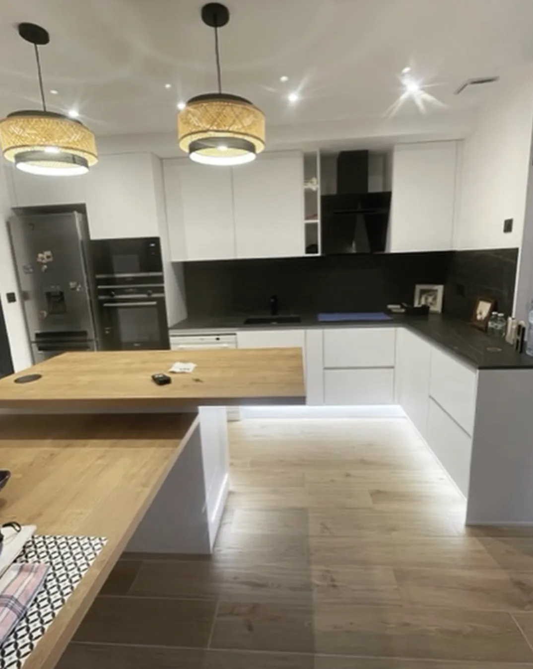 Cuisine moderne avec plan de travail en bois clair, armoires blanches, électroménagers noirs et éclairage suspendu.