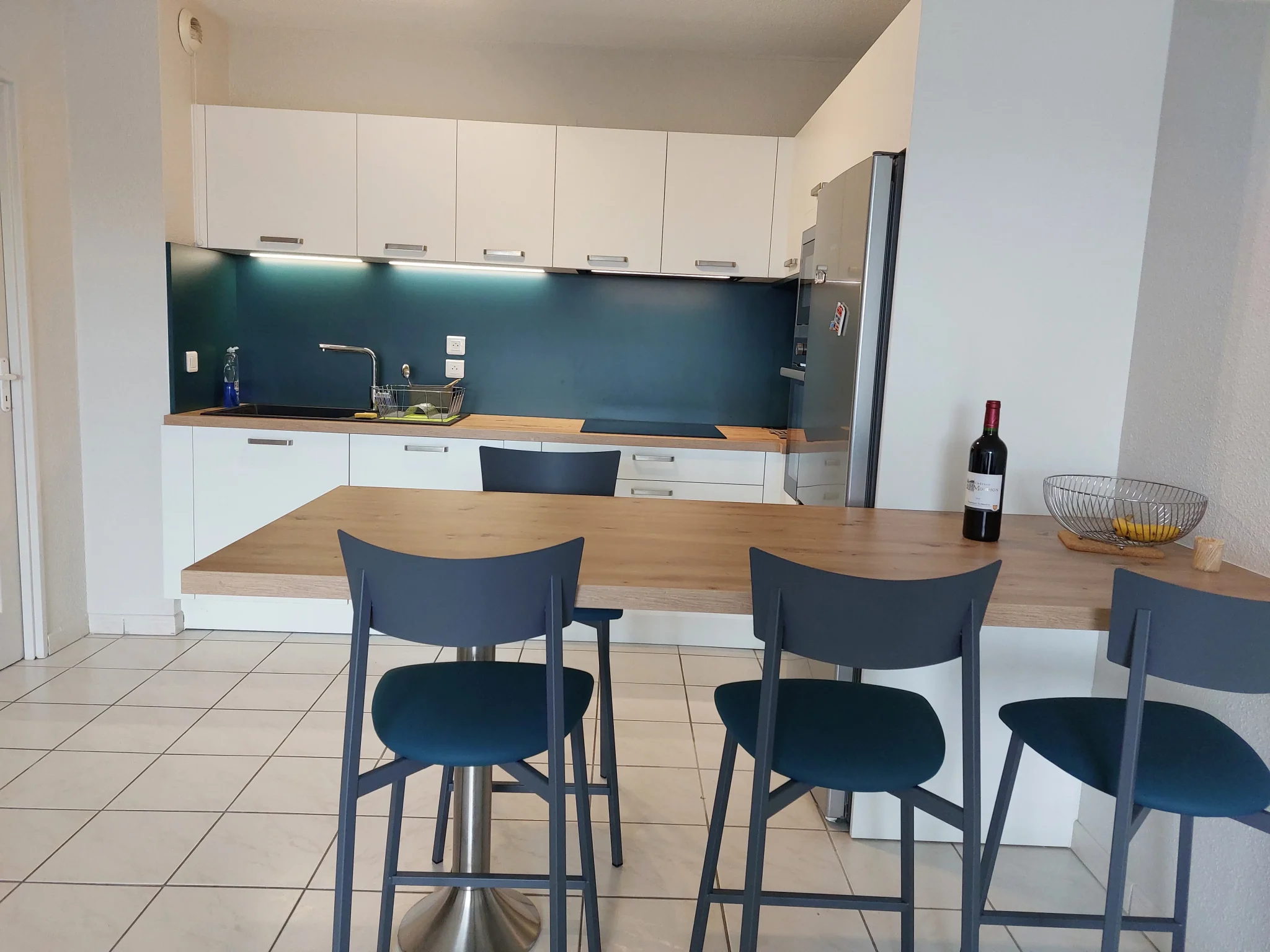 Cuisine moderne avec comptoir en bois, trois chaises bleues, évier, réfrigérateur inox, et bouteille de vin rouge sur le côté.