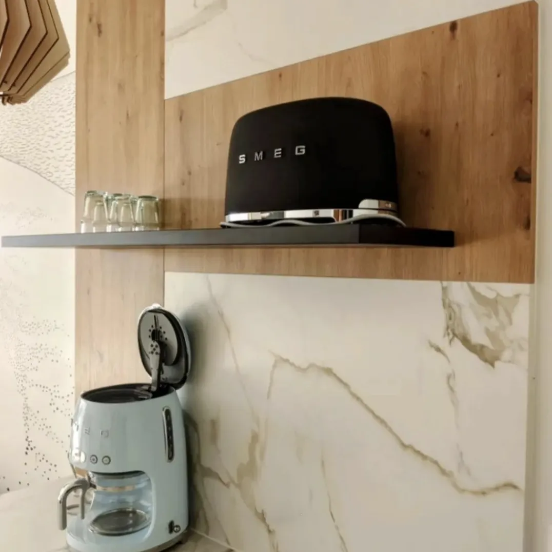 Grille-pain noir SMEG sur une étagère noire avec plusieurs petits verres à côté, et machine à café bleu clair avec couvercle ouvert sous l'étagère contre un mur en marbre et un panneau en bois.