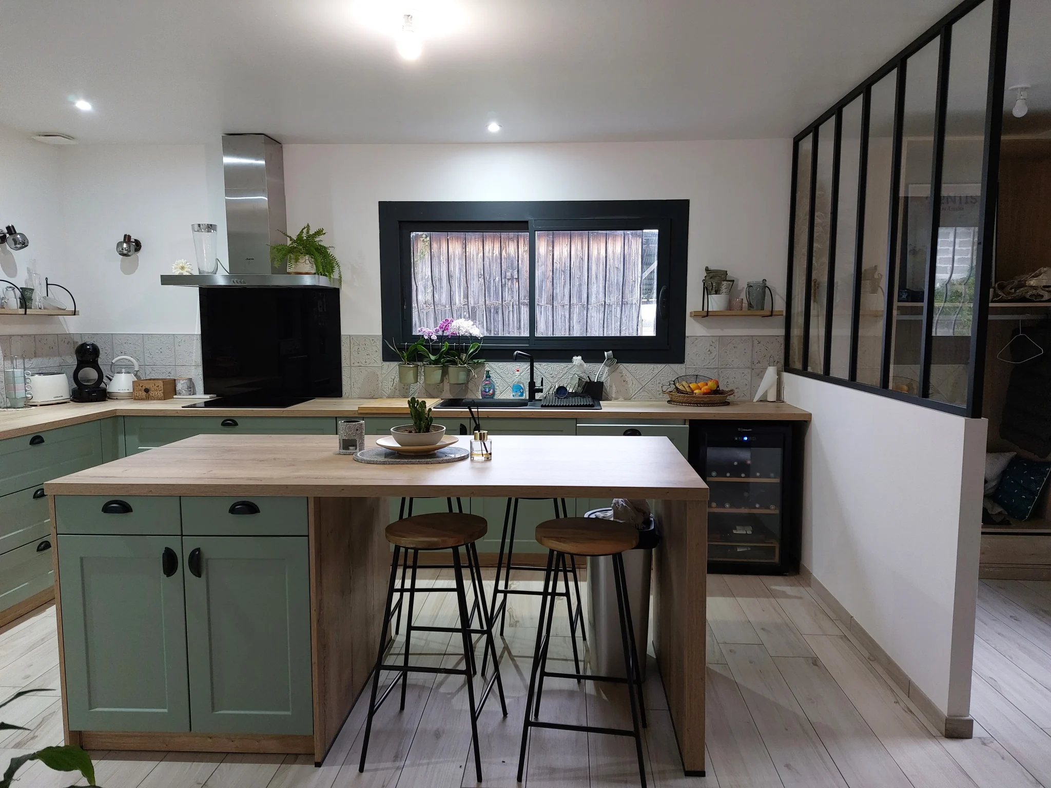 Cuisine moderne avec meubles verts, plan de travail en bois clair, îlot central avec tabourets, et fenêtre noire donnant sur un mur en bois.