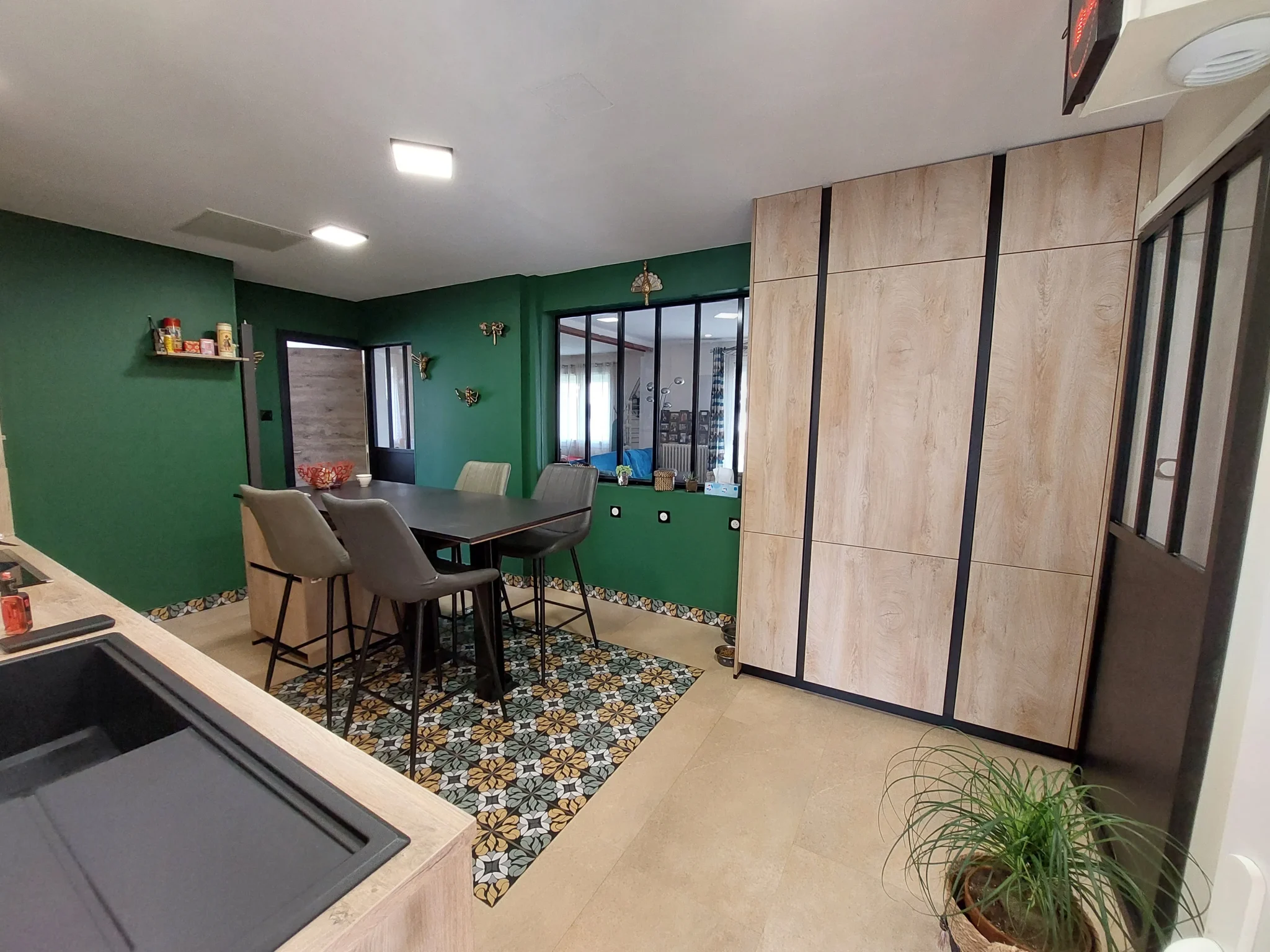 Cuisine moderne avec murs verts, table haute noire entourée de chaises grises, sol en carrelage beige avec tapis coloré, grand placard en bois clair, et plante en pot.