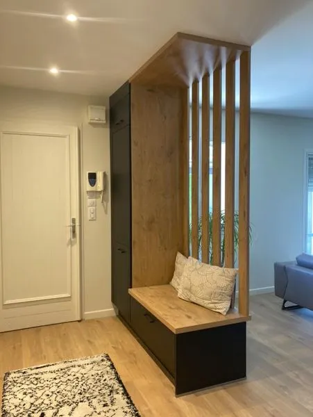 Entrée intérieure avec un banc en bois clair et noir avec un coussin, des lattes verticales en bois en arrière-plan, un tapis blanc à motifs noirs, une porte blanche et un interphone mural.