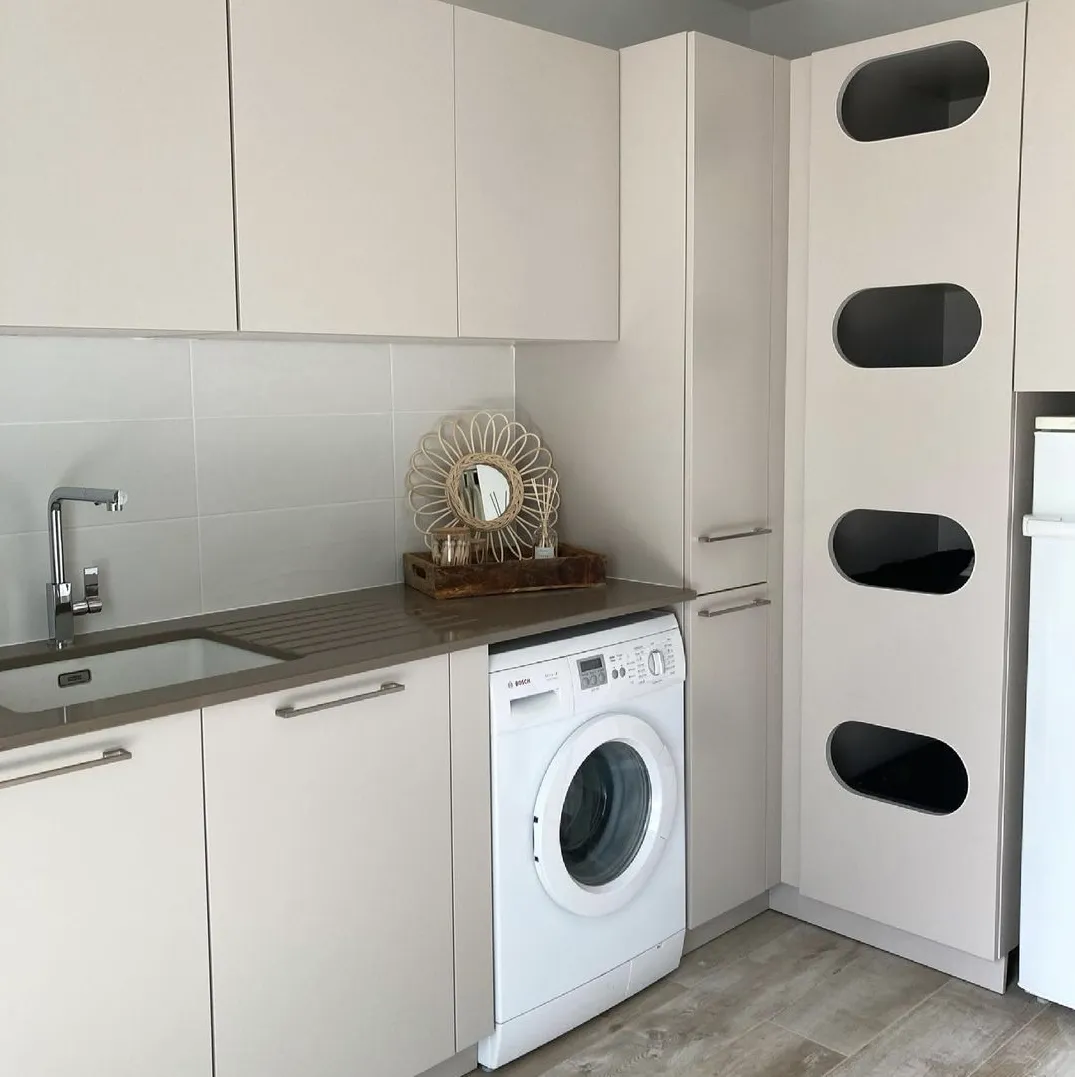 Buanderie moderne avec lave-linge blanc intégré, évier, et placards beiges avec plan de travail gris.