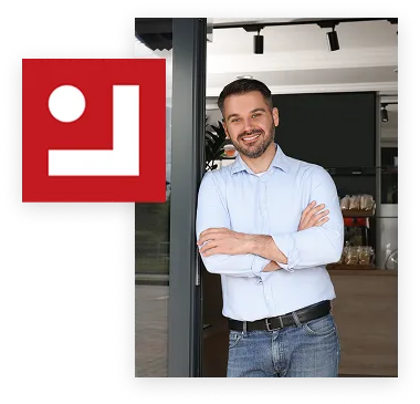 Homme souriant en chemise bleue à manches longues et jean, bras croisés, debout devant une porte vitrée dans un espace moderne.