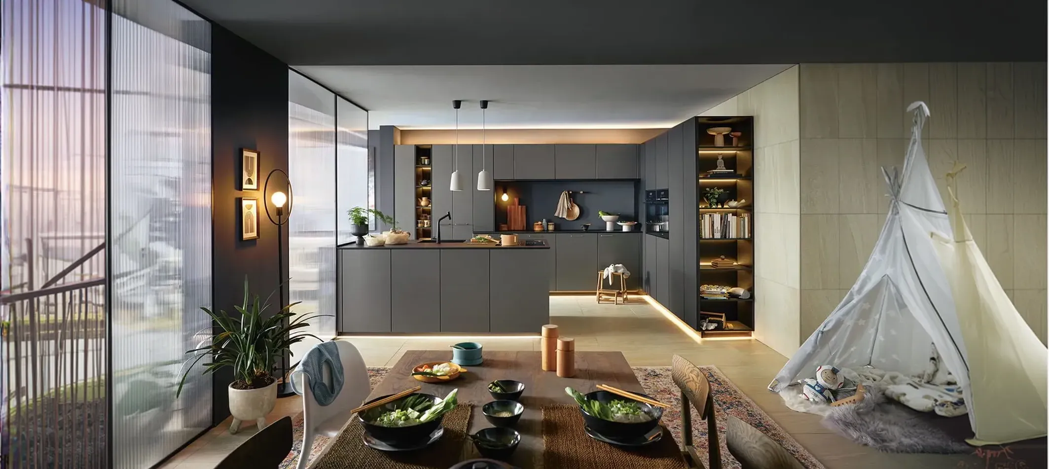 Cuisine moderne avec meubles gris, îlot central, salle à manger avec bols et baguettes, et tente de jeu pour enfants près d'une grande fenêtre.