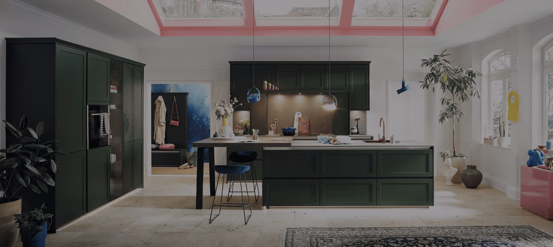 Cuisine moderne avec îlot central vert foncé, chaises, plantes et grandes fenêtres sous un plafond en verre.
