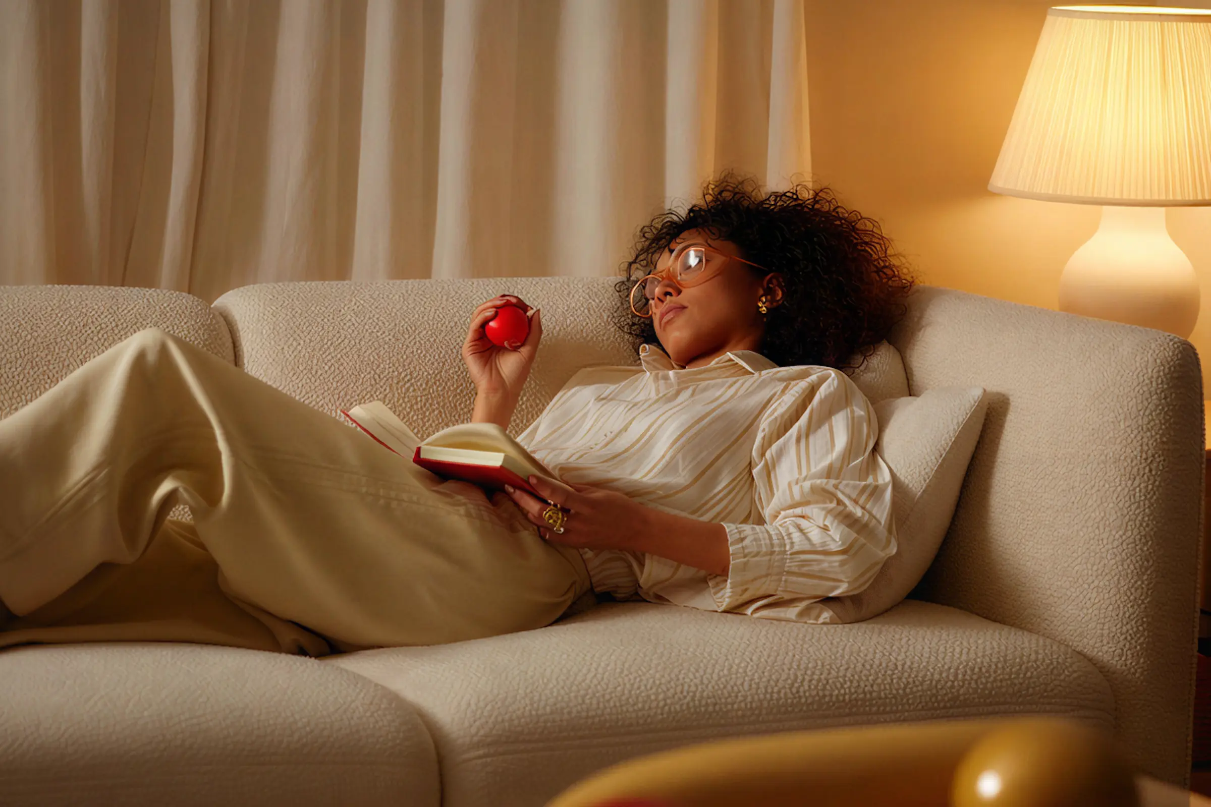 Woman lounging on a beige couch holding a red stress ball and a book, with a lit lamp in the background.
