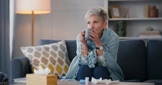 Older woman wrapped in a blanket on a couch holding a mug, tissues and medicine on the table