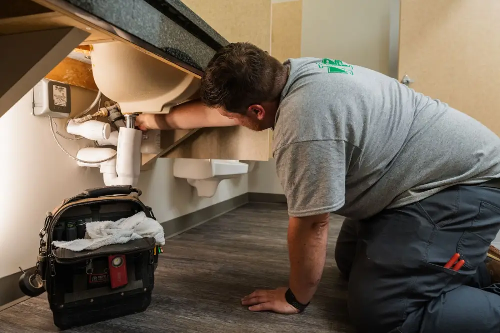 Korrect plumber kneeling to work on a bathroom sink drain trap, tool bag beside him