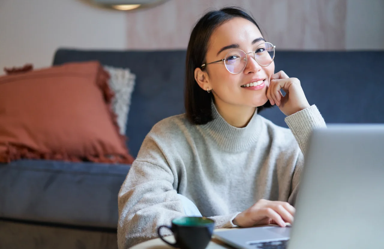Smiling woman in orange sweater using tablet with stylus while sitting at a table with documents and notebook.