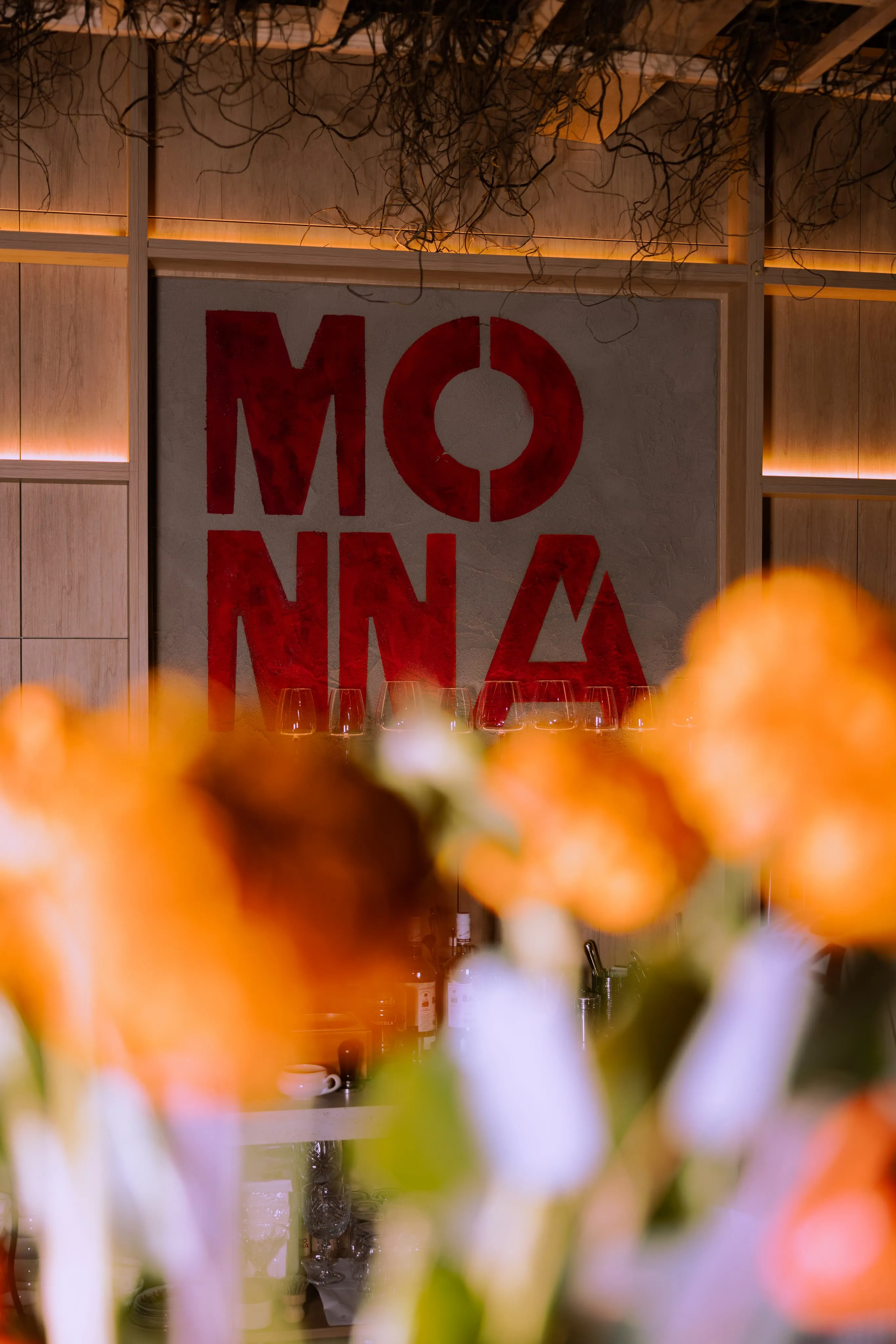 Red block letters spelling 'MONNA' on a gray wall behind a bar counter with blurred orange flowers in the foreground.