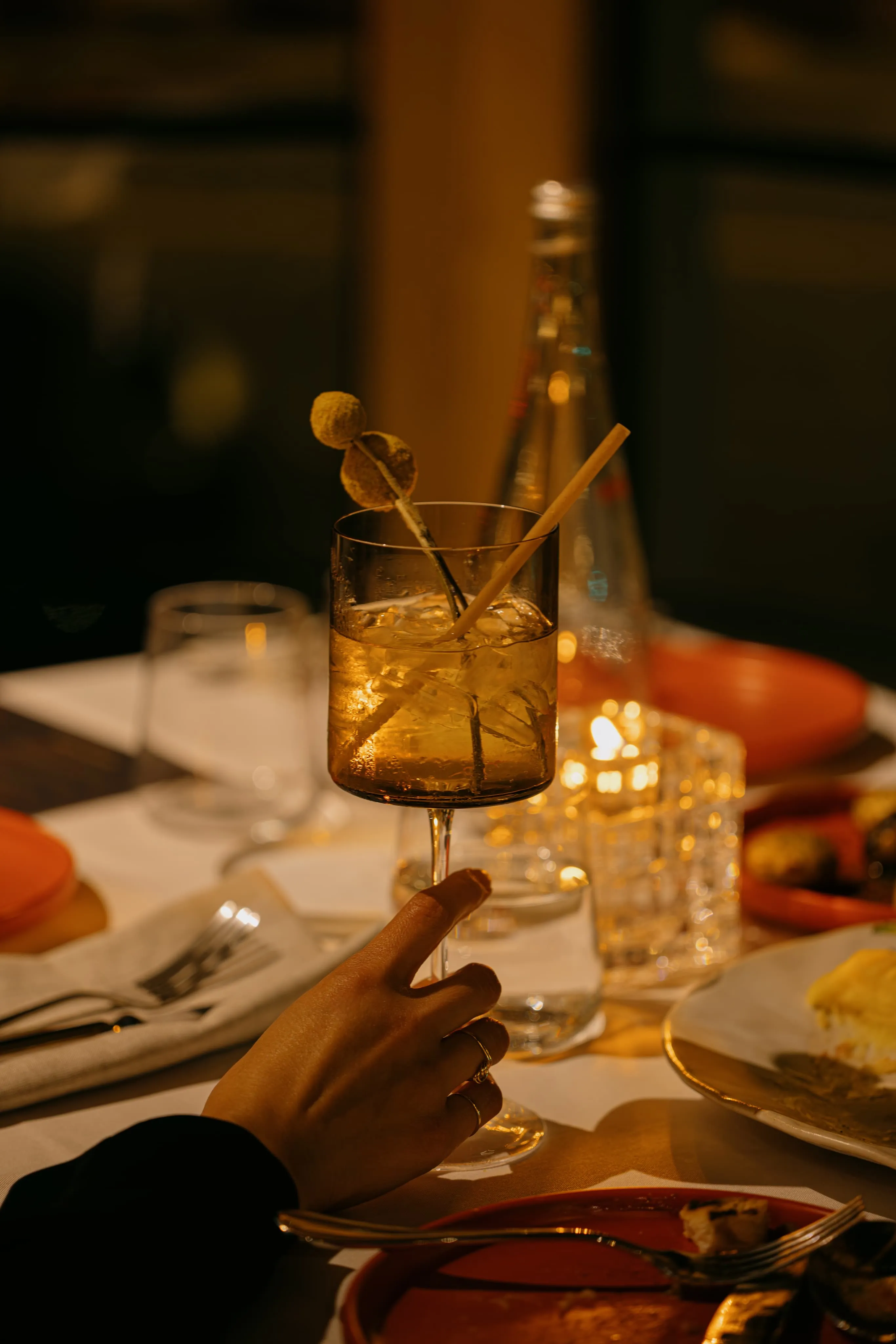 Hand holding a stemmed glass with a drink, ice, a straw, and dried decorative flowers in a dimly lit dining setting.