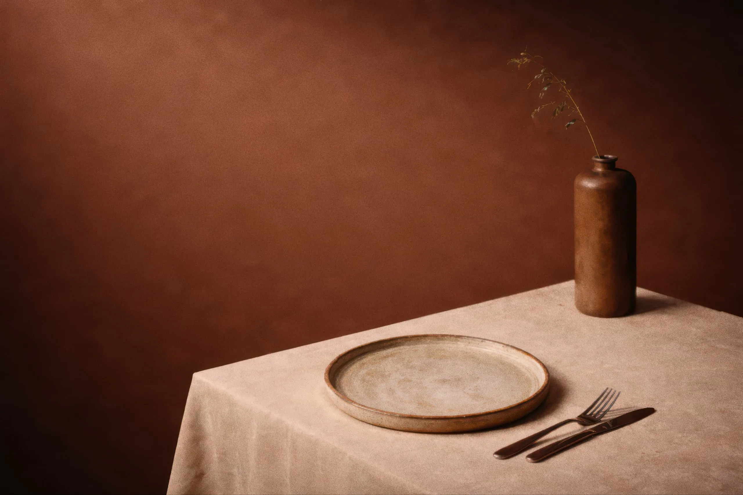 Empty ceramic plate with fork and knife on a beige tablecloth beside a brown vase with a single dried branch.