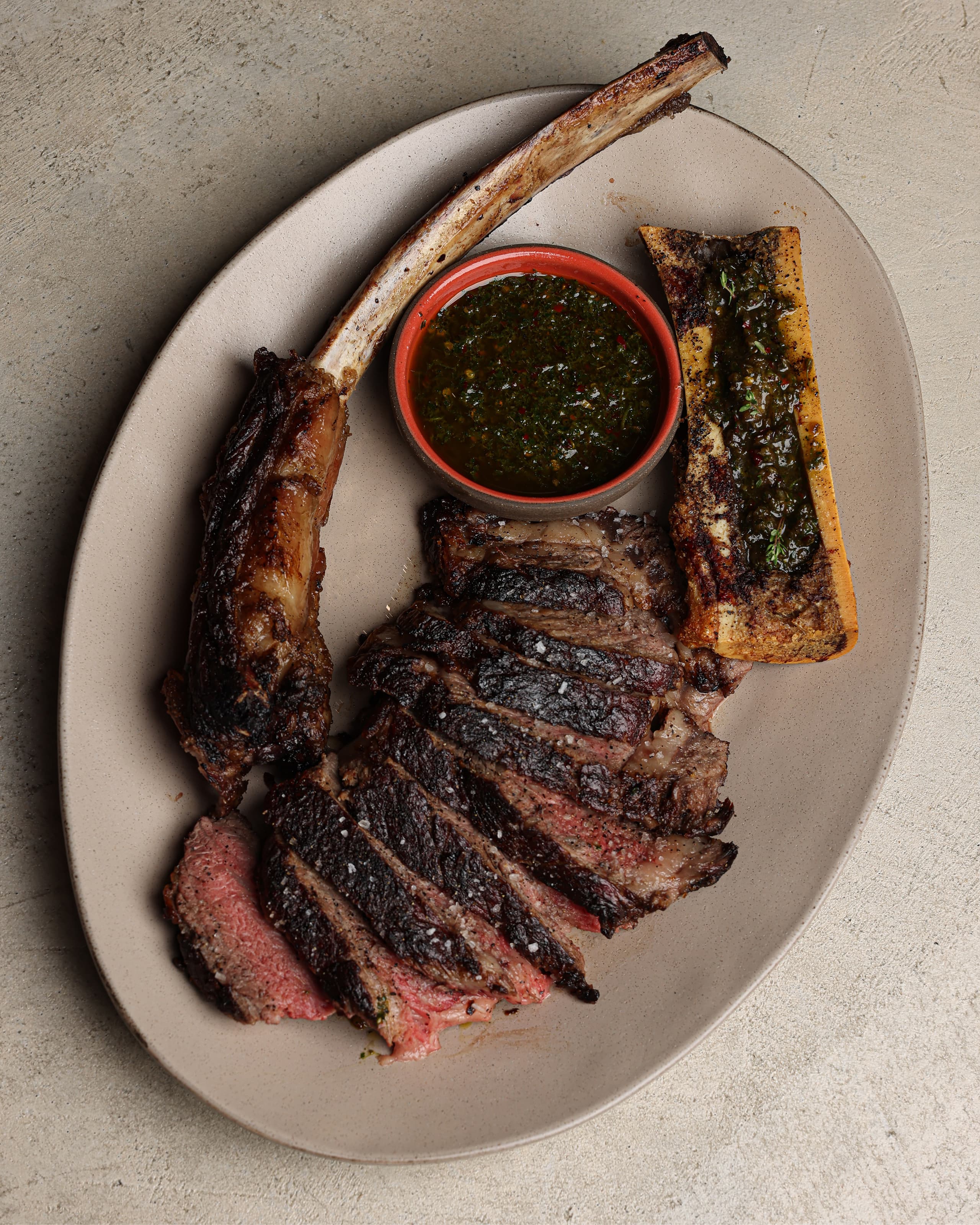 Sliced grilled tomahawk steak with a bone marrow piece and a bowl of green herb sauce on a beige plate.
