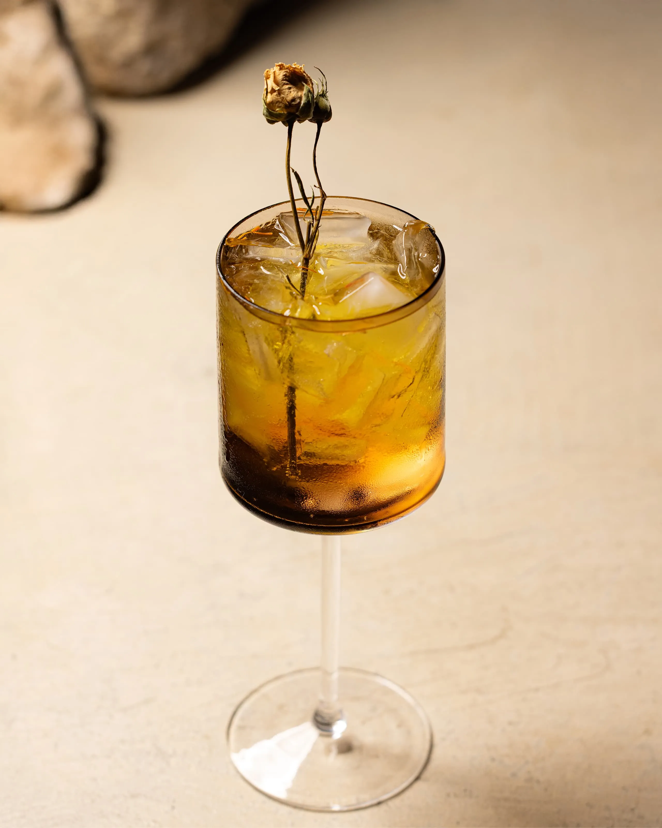 Tall stemmed glass filled with an amber iced drink garnished with dried flowers on a beige surface.
