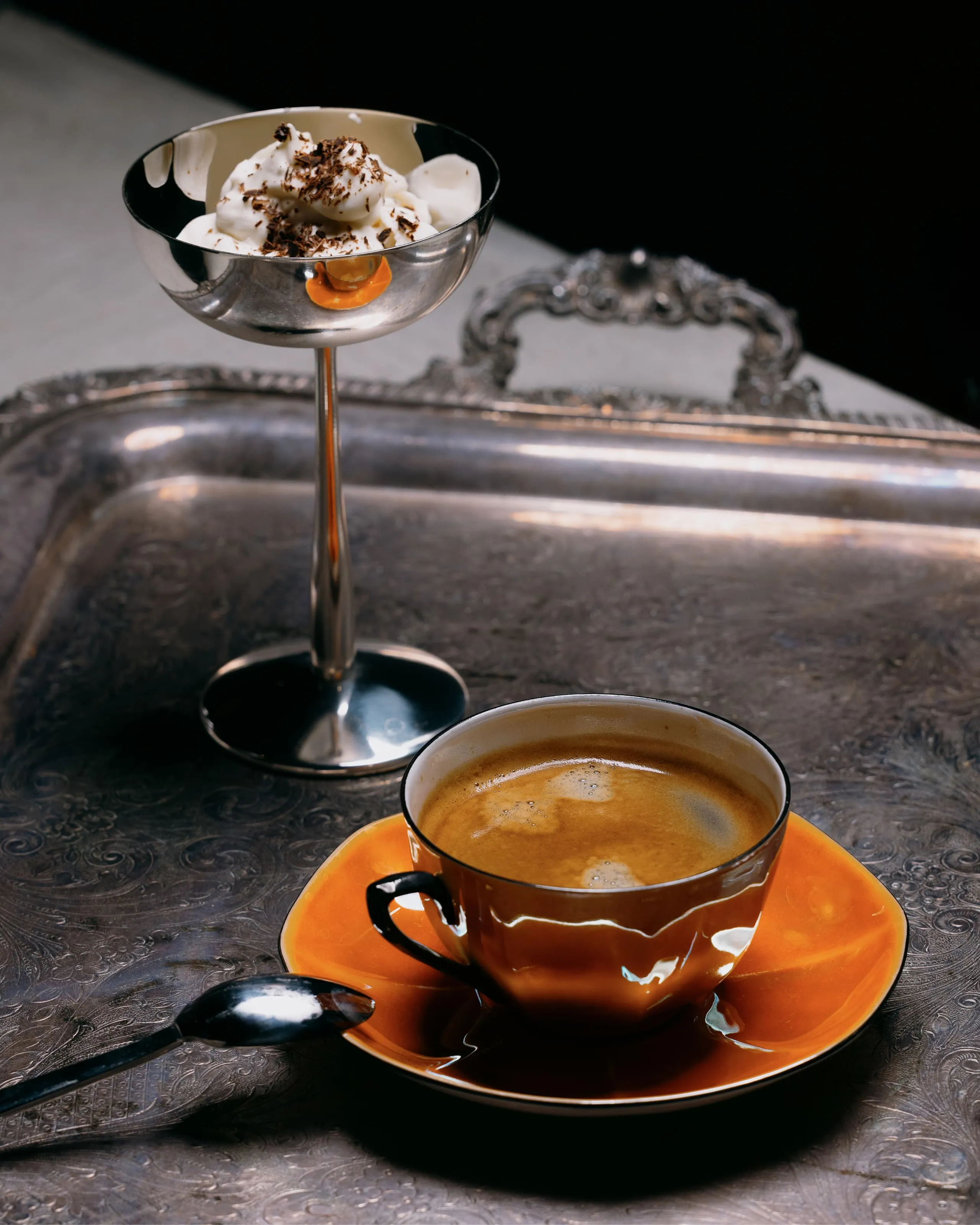 Orange cup of coffee with saucer and spoon on an ornate silver tray next to a silver dessert dish with whipped cream and chocolate shavings.
