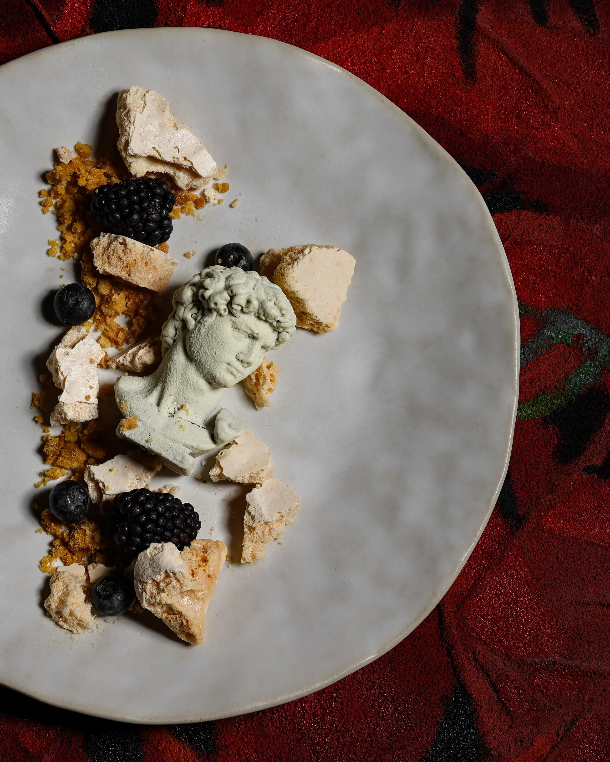 White plate with a small bust sculpture surrounded by crumbled meringue pieces, blackberries, and blueberries on a red textured surface.