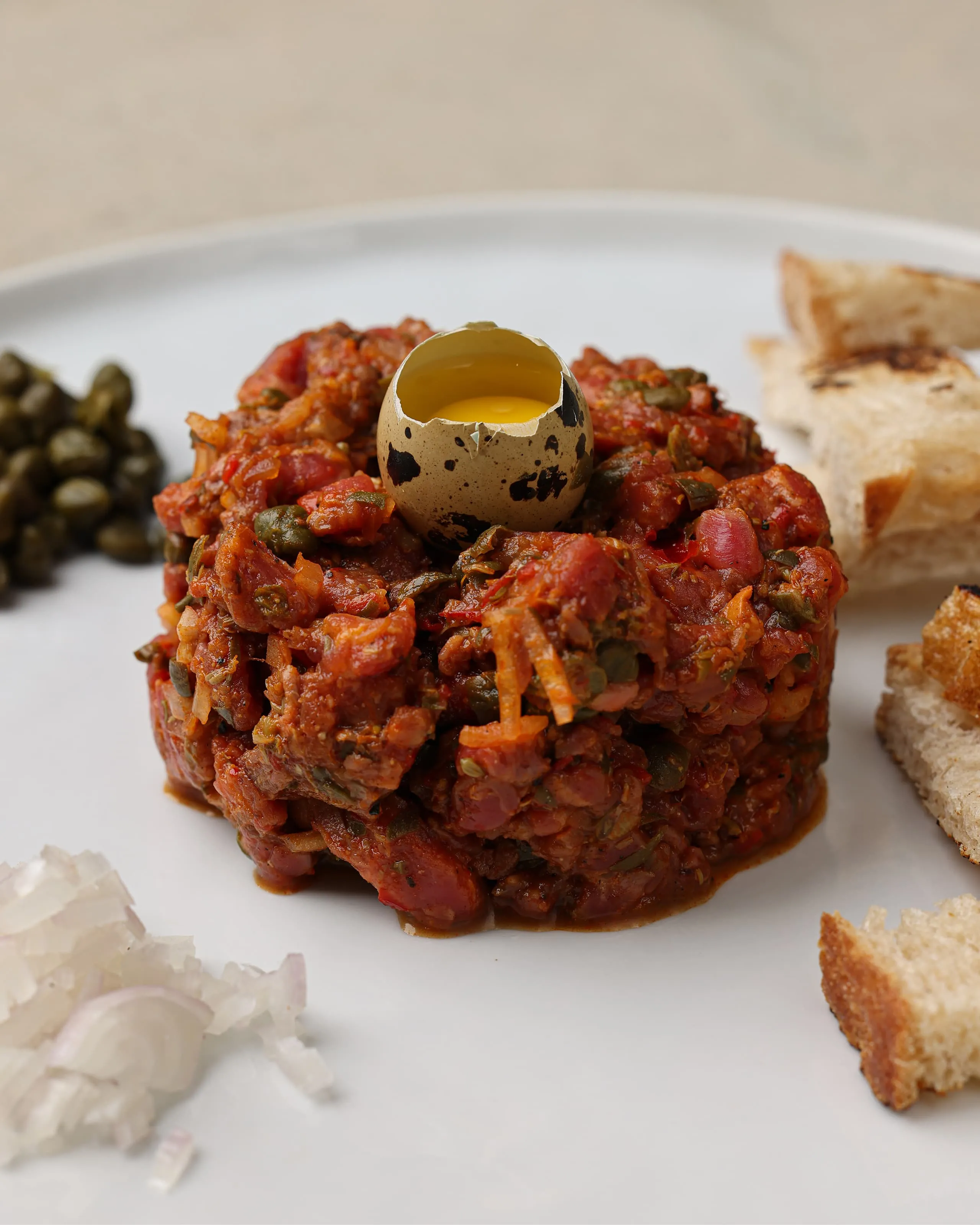 Plate with a mound of finely chopped tartare-like mixture topped with a quail egg yolk in a speckled shell, accompanied by toasted bread pieces, chopped onions, and capers.