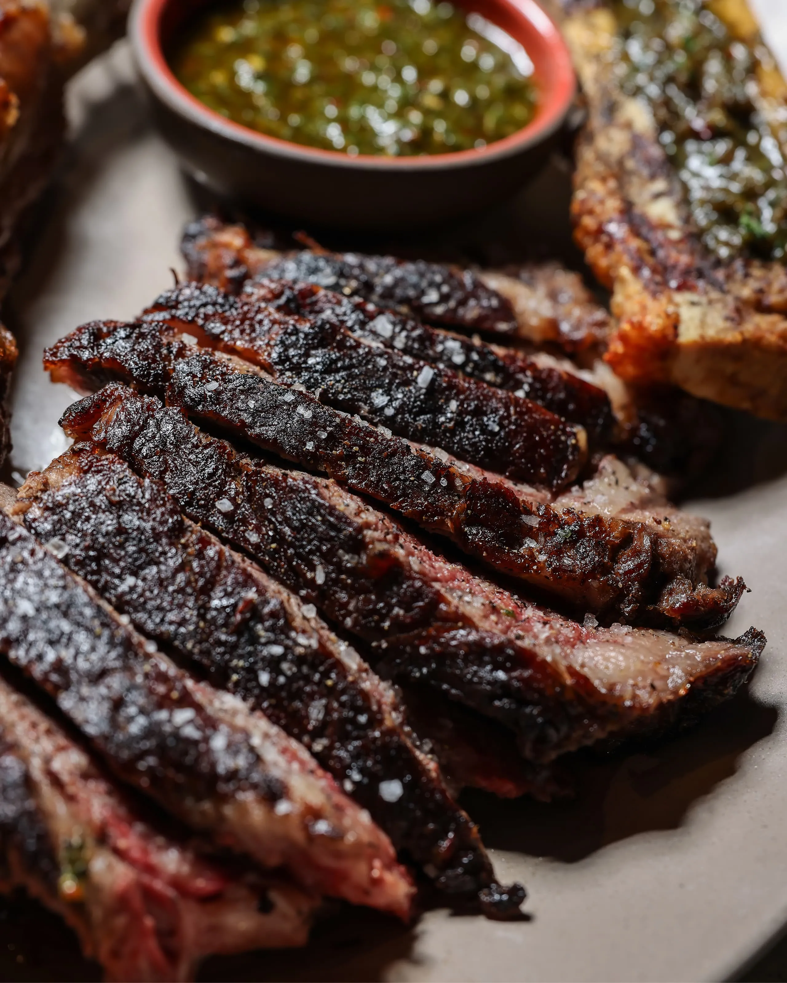 Close-up of sliced smoked brisket with coarse salt on a plate, accompanied by a green sauce in a small bowl and a piece of grilled bread.