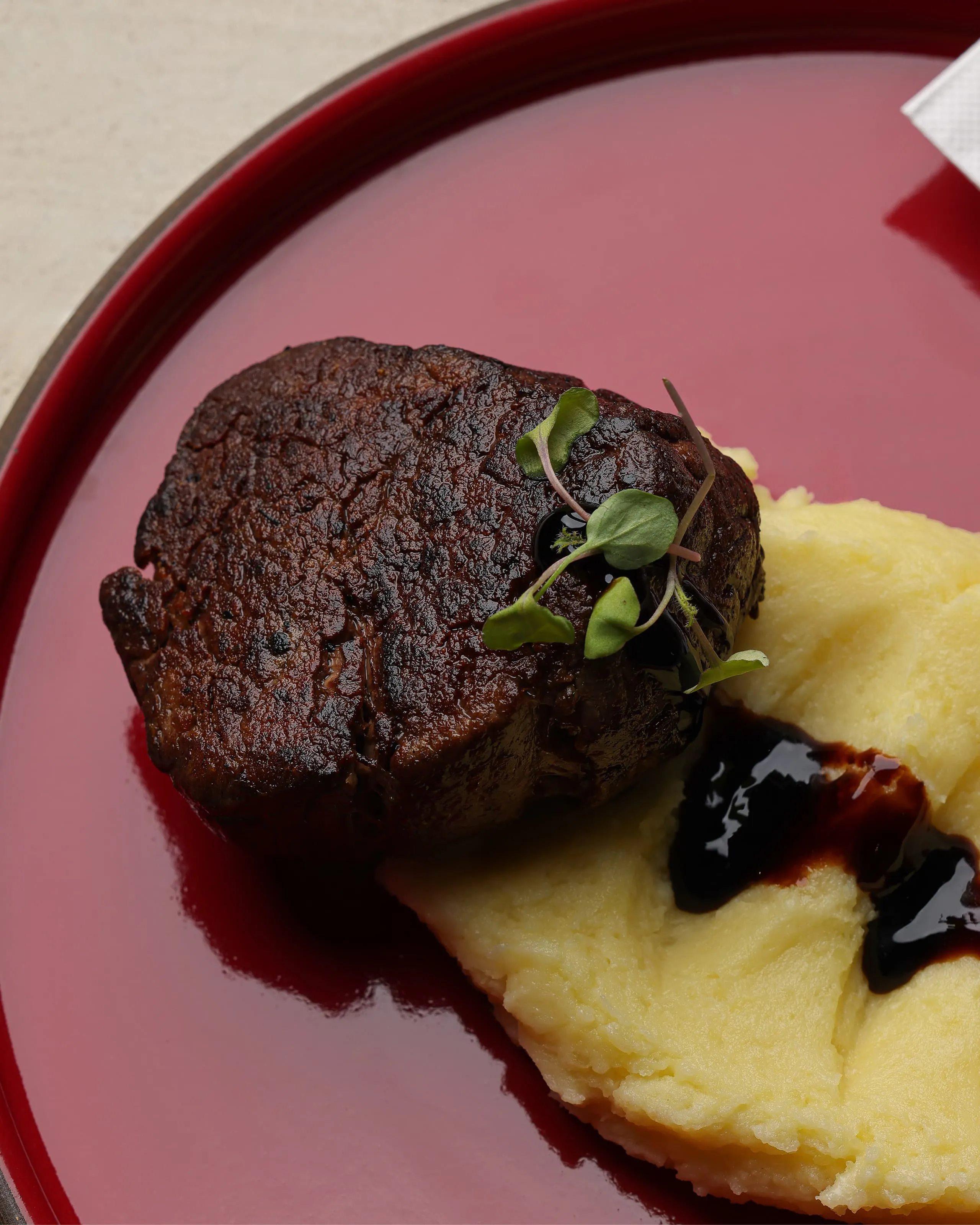 Seared steak filet garnished with microgreens served with creamy mashed potatoes and dark sauce on a red plate.