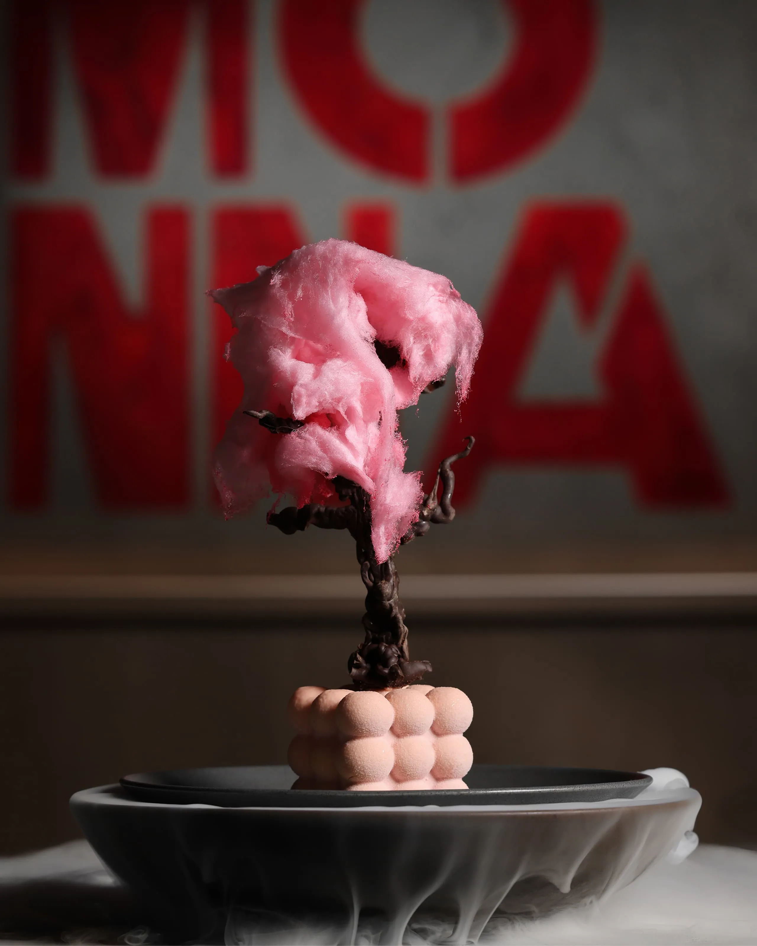 Artistic dessert with pink cotton candy atop a chocolate tree on a pale pink cube, placed on a plate with smoke effect.