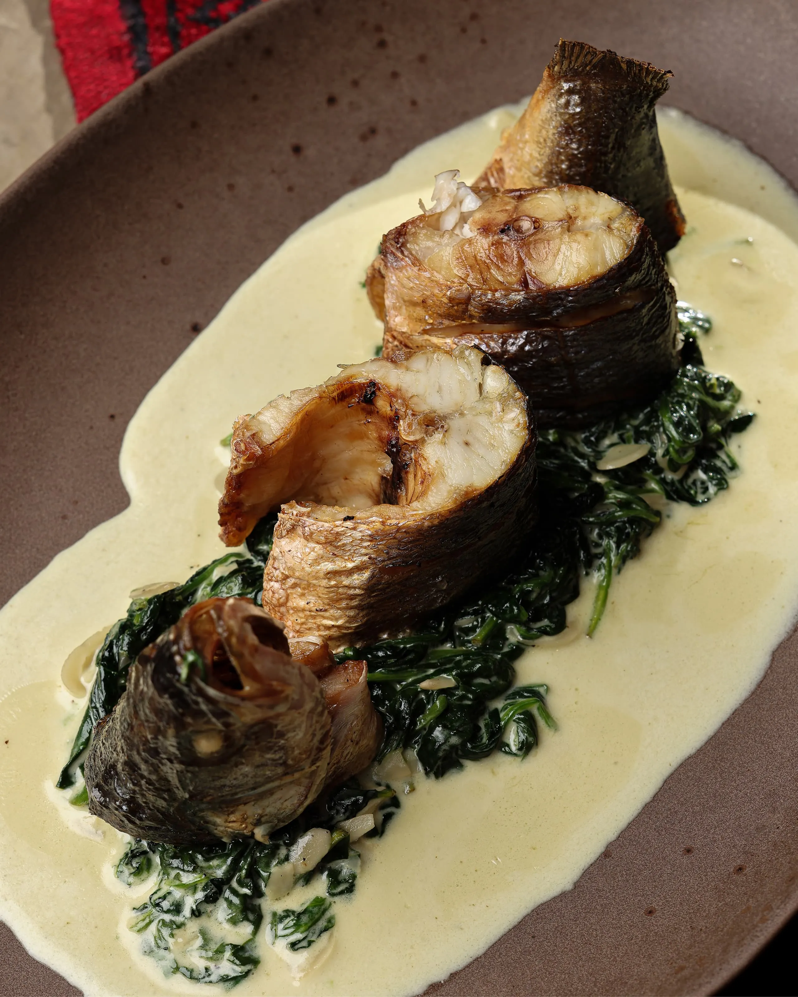 Grilled fish pieces served on a bed of creamed spinach with a light creamy sauce on a brown plate.