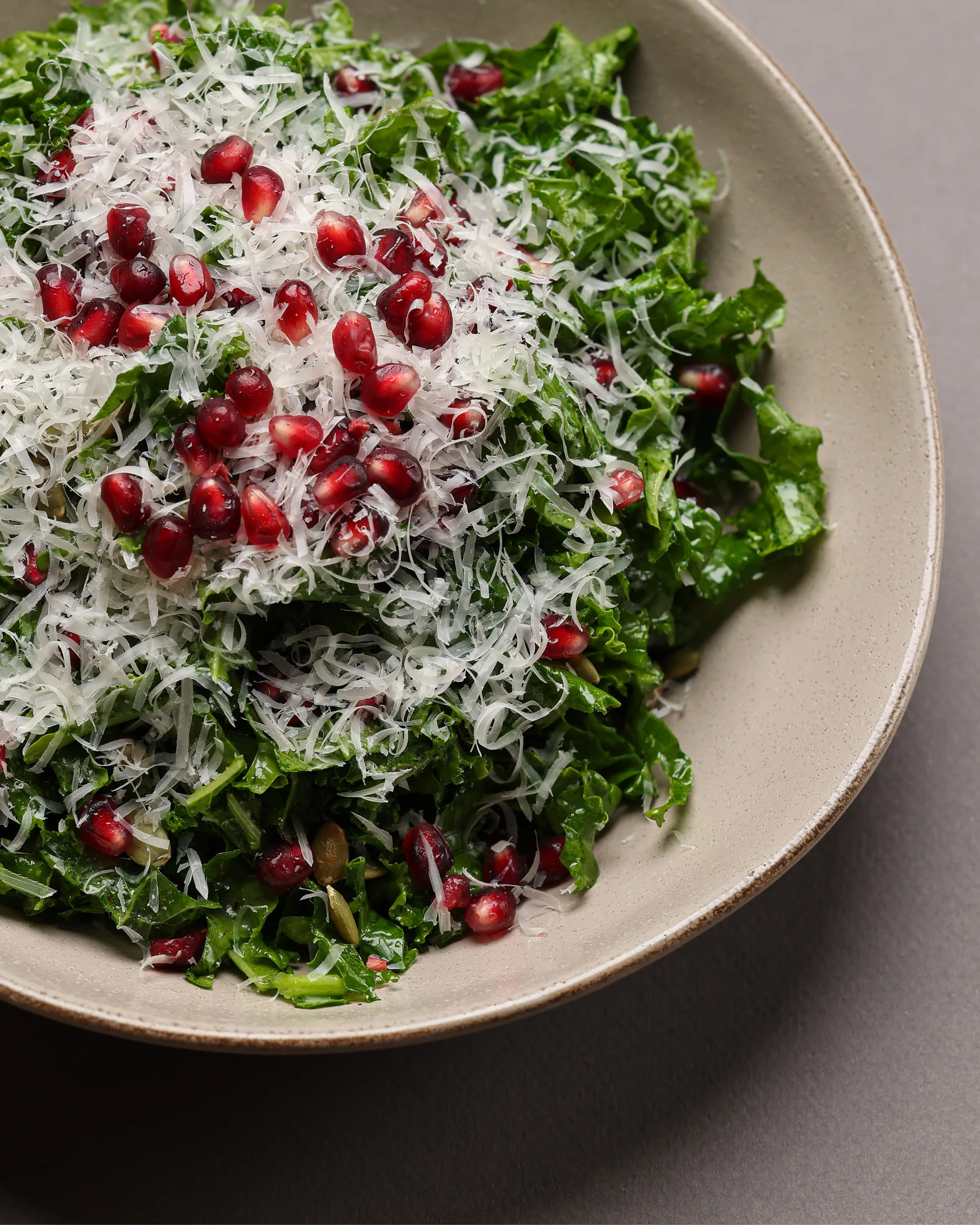 Bowl of fresh kale salad topped with grated cheese and scattered pomegranate seeds.
