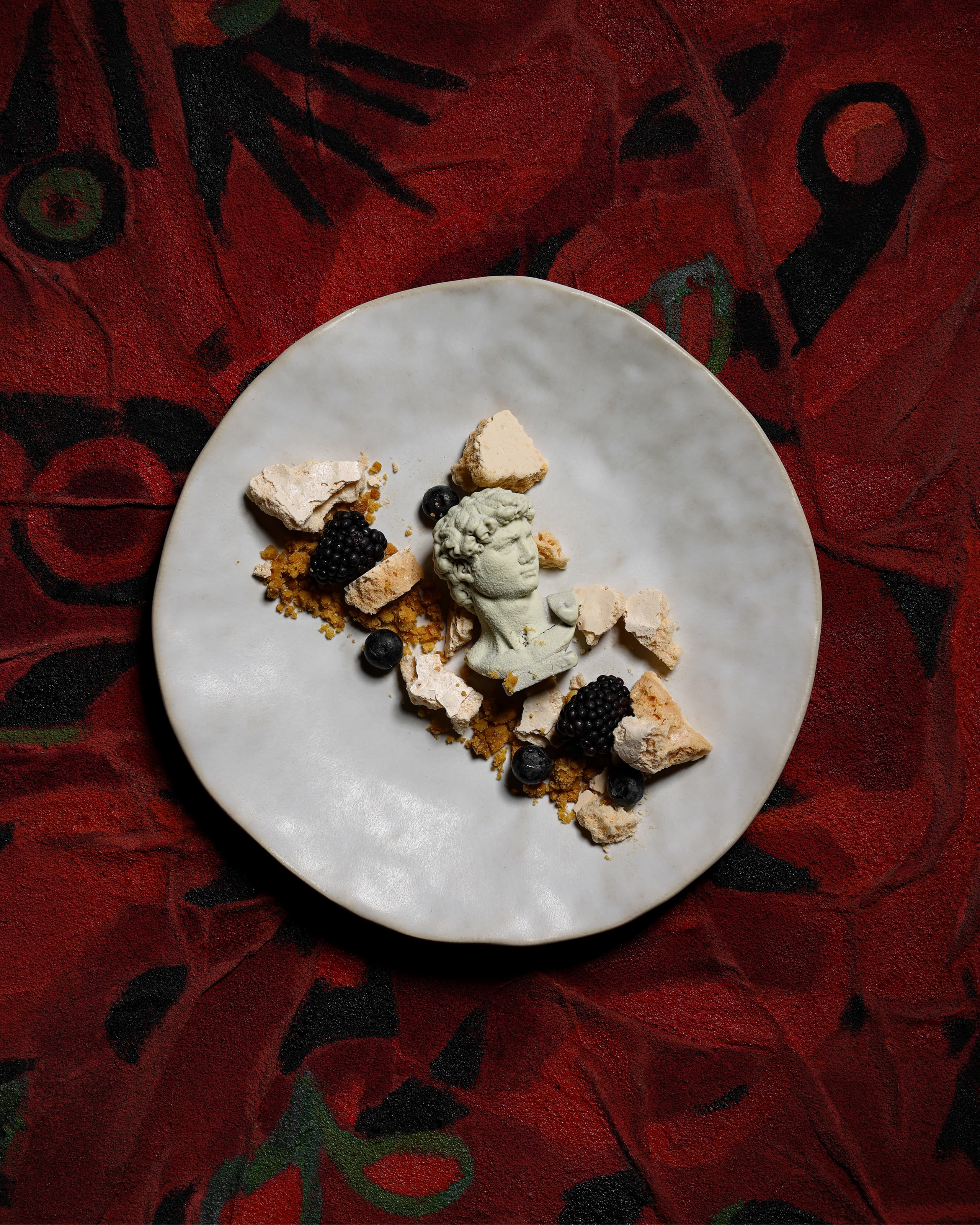 White ceramic plate with dessert featuring a small bust sculpture, crumbled meringue, blackberries, blueberries, and crumbs on a red patterned background.