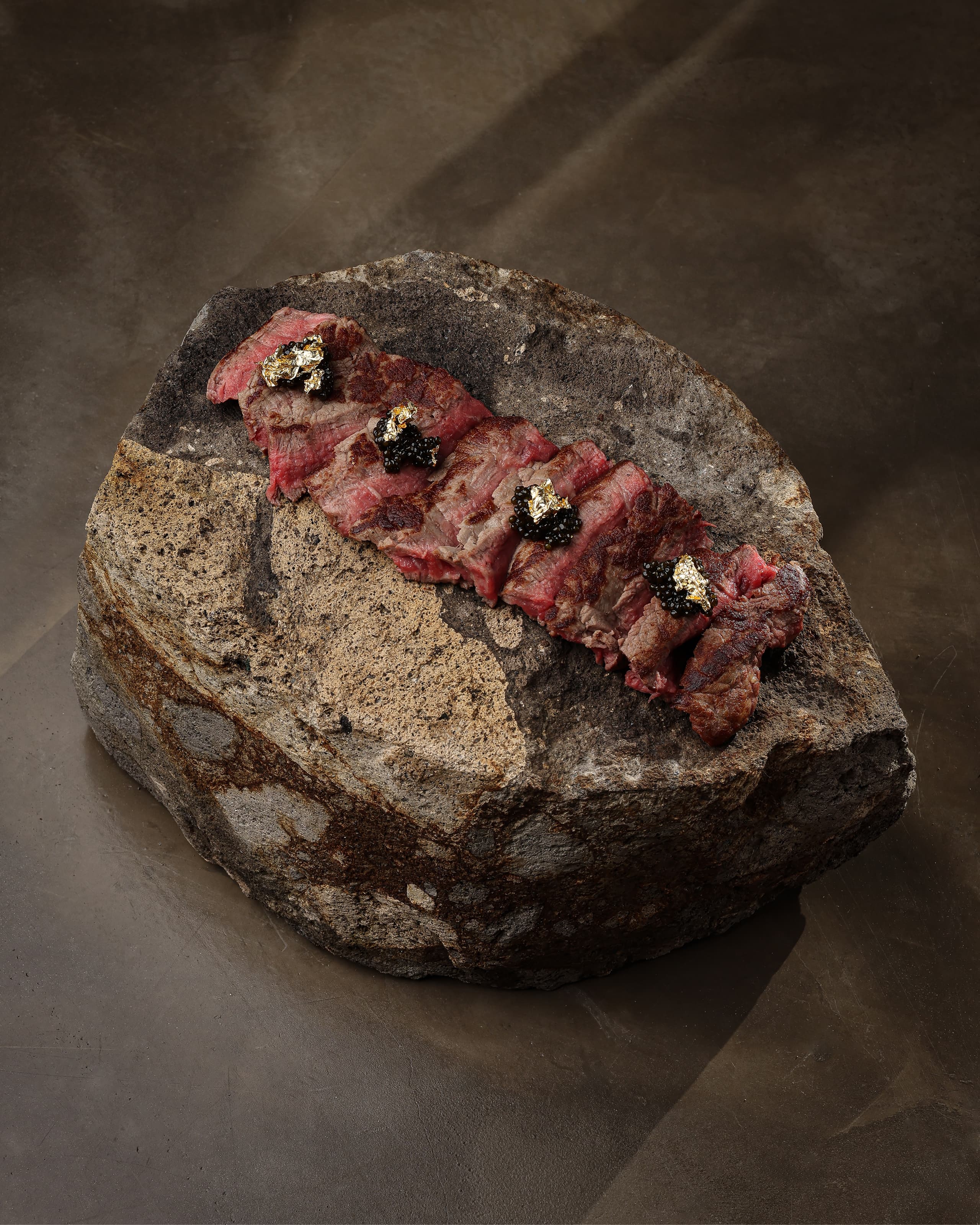 Sliced rare steak topped with black caviar and gold leaf, served on a large rough stone slab.