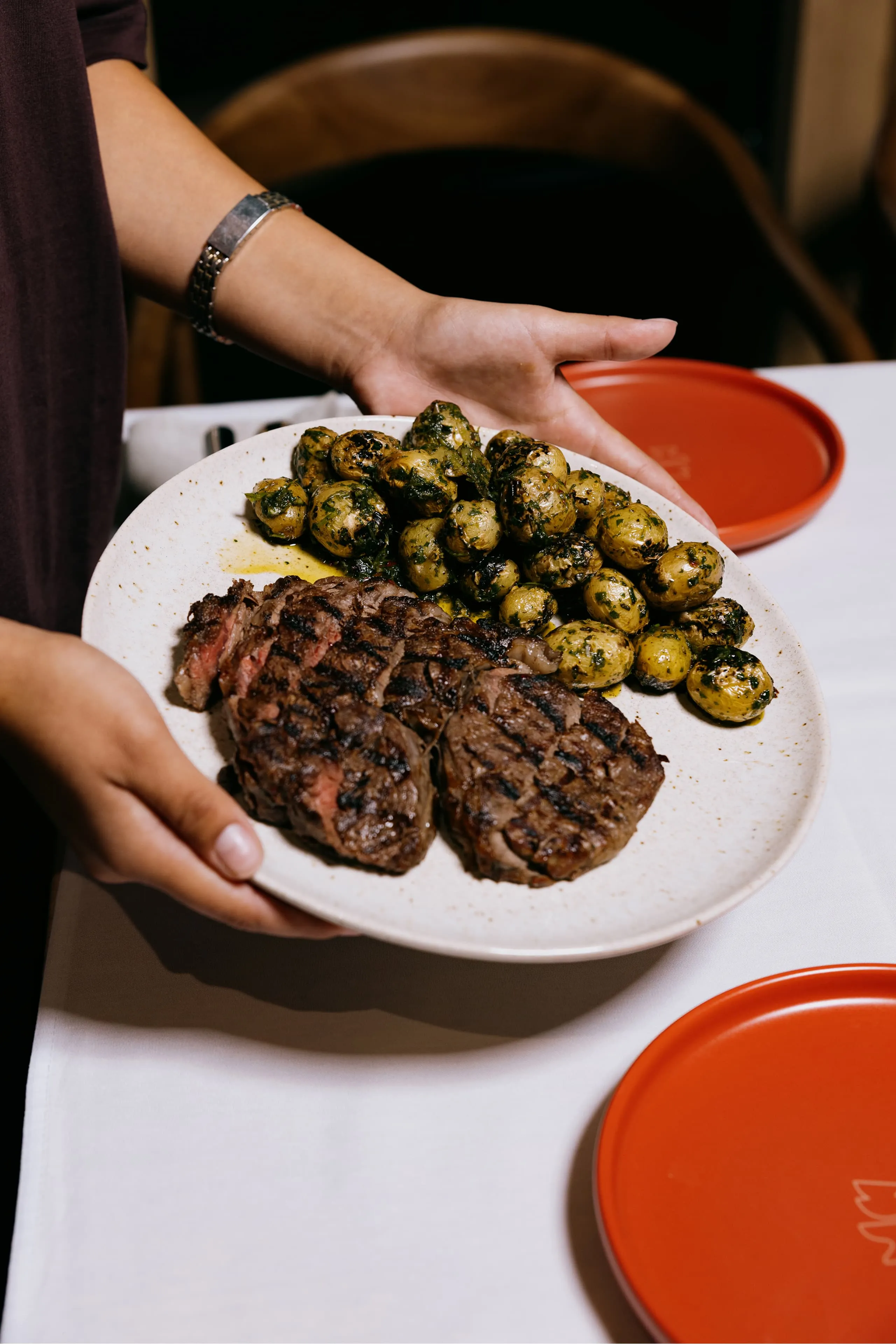 Two grilled steaks and a serving of herb-seasoned baby potatoes on a white plate held by a person.