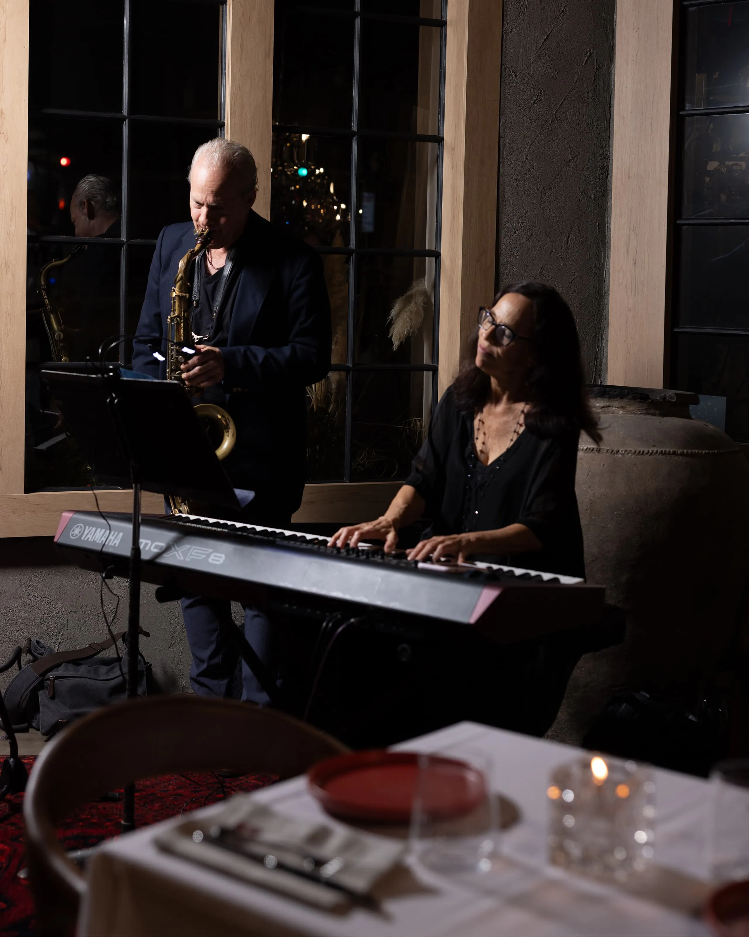 Man playing saxophone and woman playing Yamaha keyboard in a dimly lit room with a dining table in the foreground.