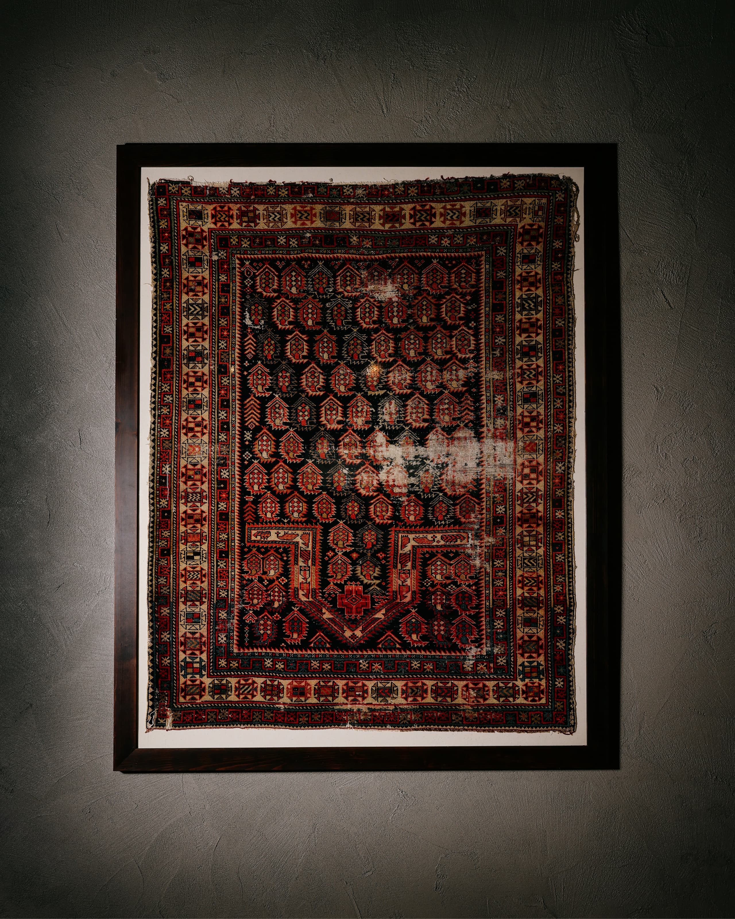 Framed vintage rug with intricate red, black, and beige geometric patterns hanging on a textured wall.