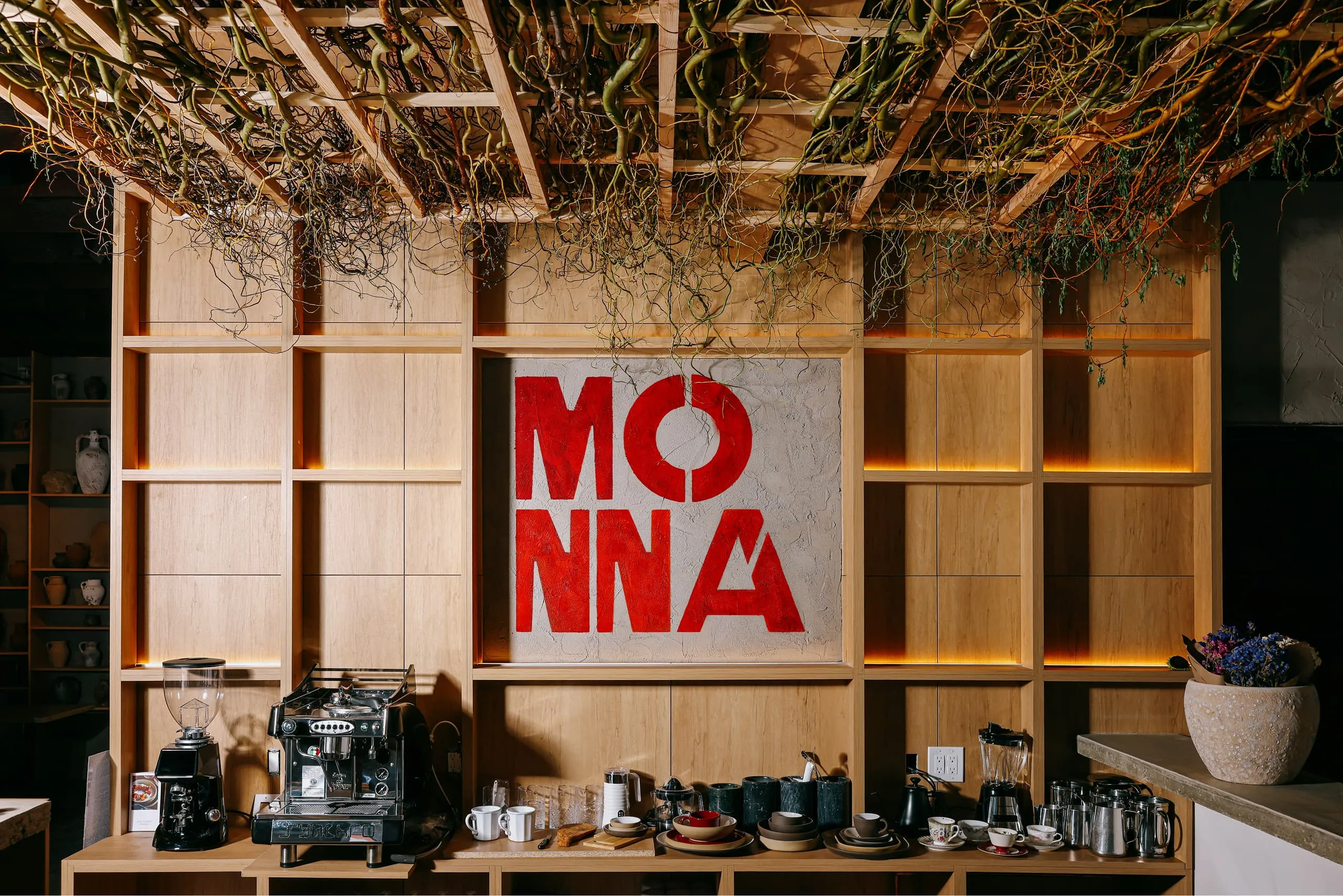 Modern coffee bar setup with a wooden shelving unit, espresso machine, coffee grinder, cups, and a red sign reading 'MONNA'.