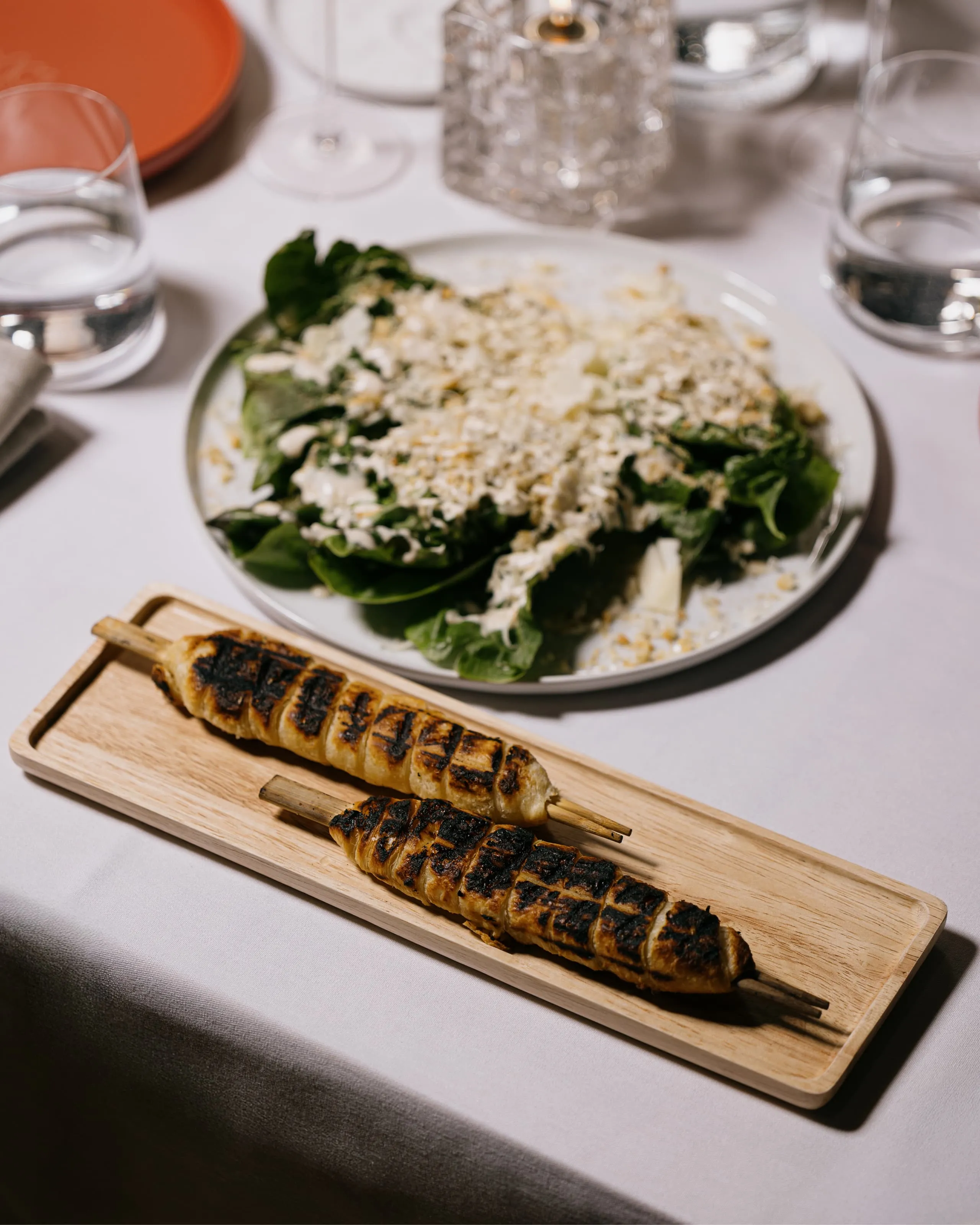Two grilled skewers with char marks served on a wooden tray, with a plate of leafy green salad topped with crumbled cheese and dressing in the background on a white tablecloth.