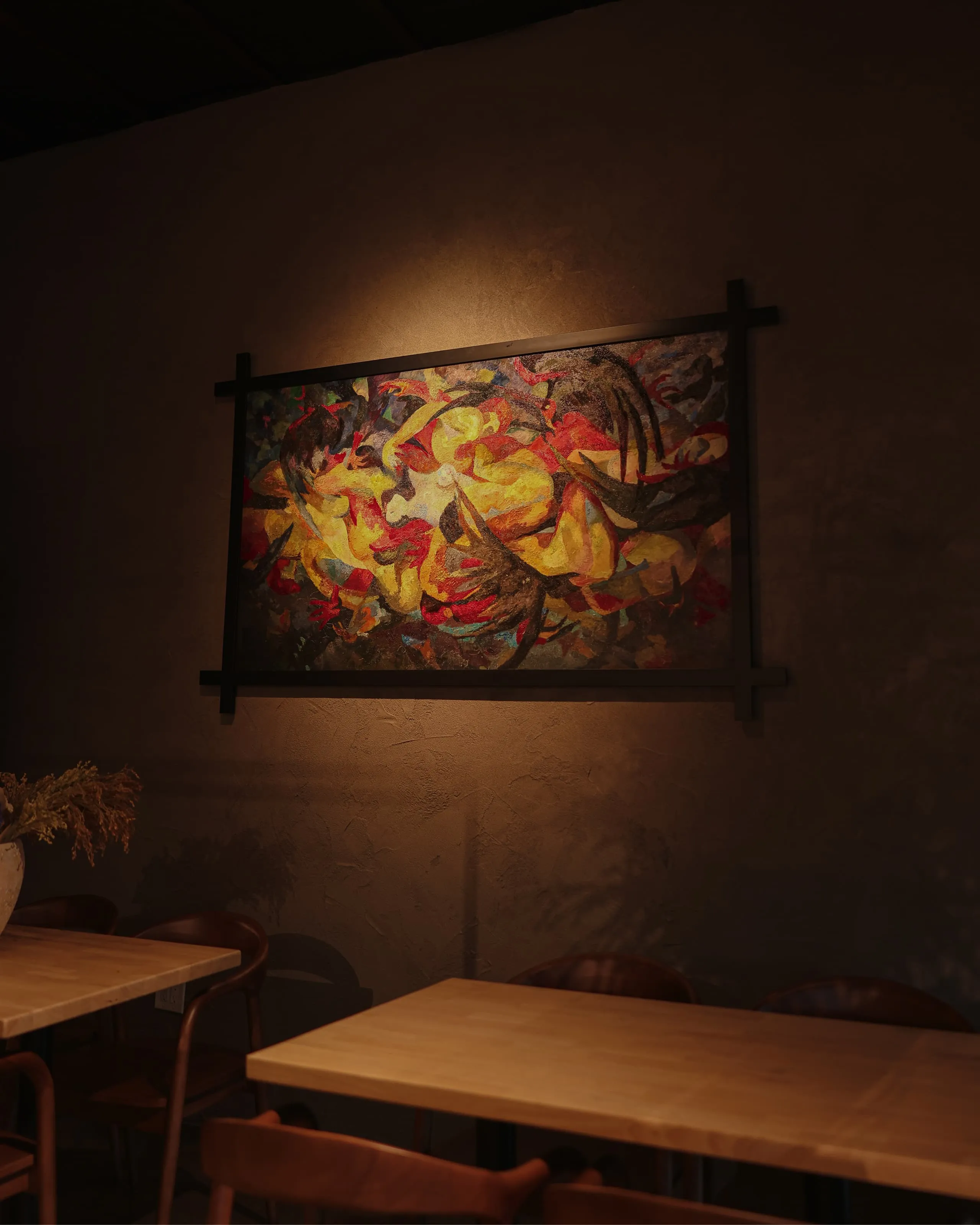 Abstract colorful painting with yellow, red, and brown tones hanging on a dark textured wall above wooden tables and chairs in a dimly lit room.