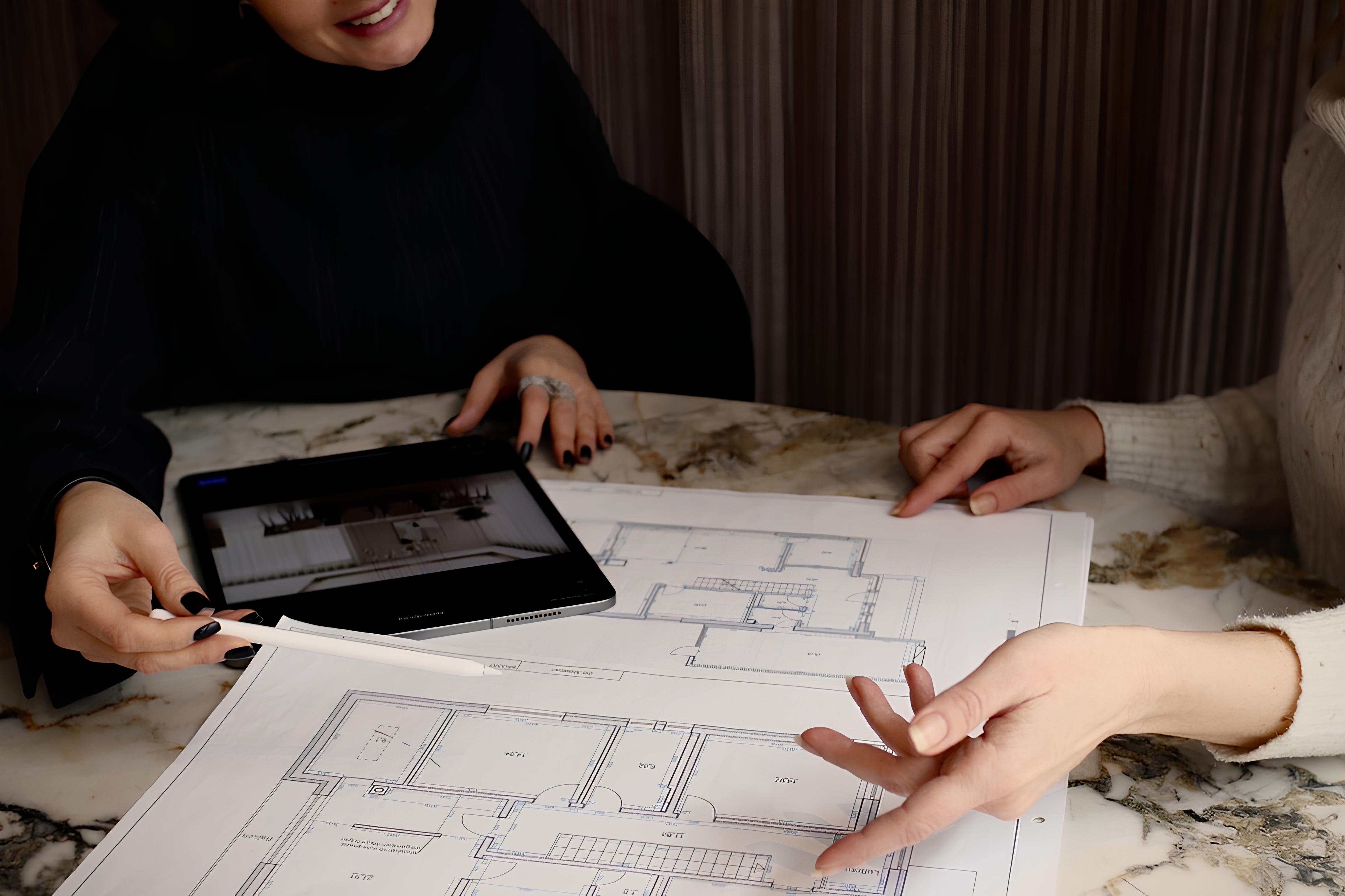 Two people reviewing architectural floor plans on a marble table with a tablet nearby.