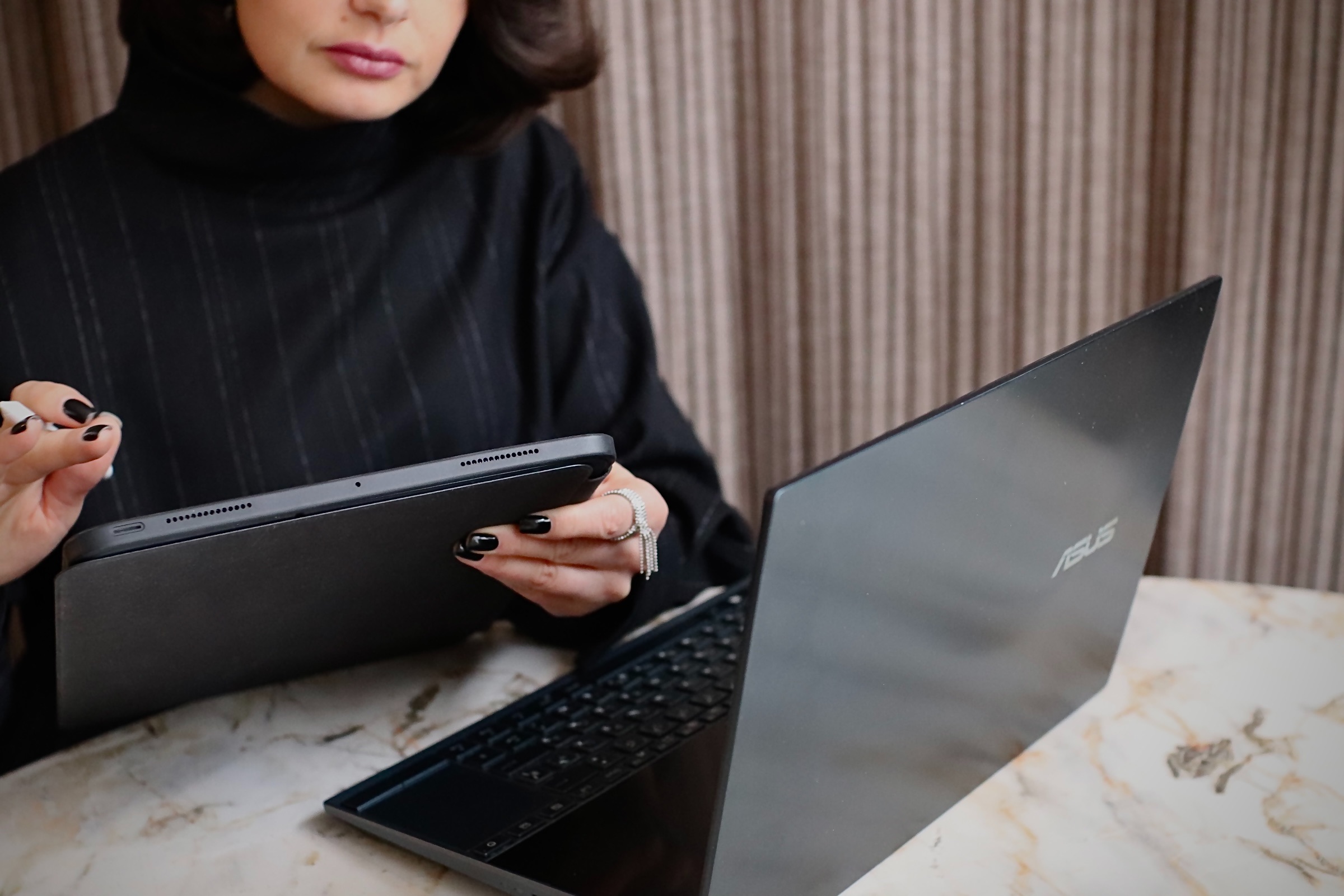 Woman in black sweater holding a tablet and using a black Asus laptop on a marble table.